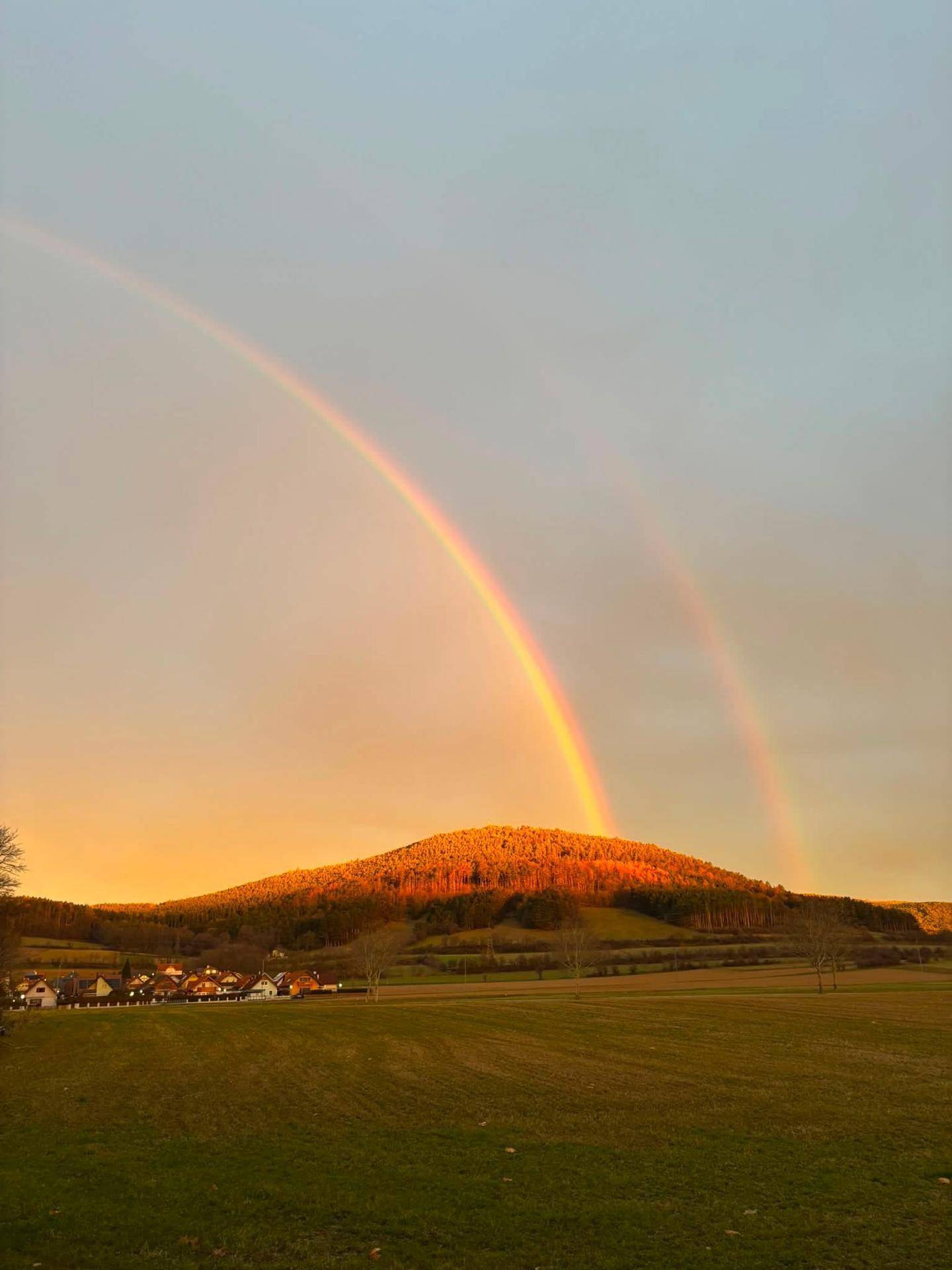 Spektakulärer doppelter Regenbogen über einer ländlichen Landschaft mit bewaldeten Hügeln.
