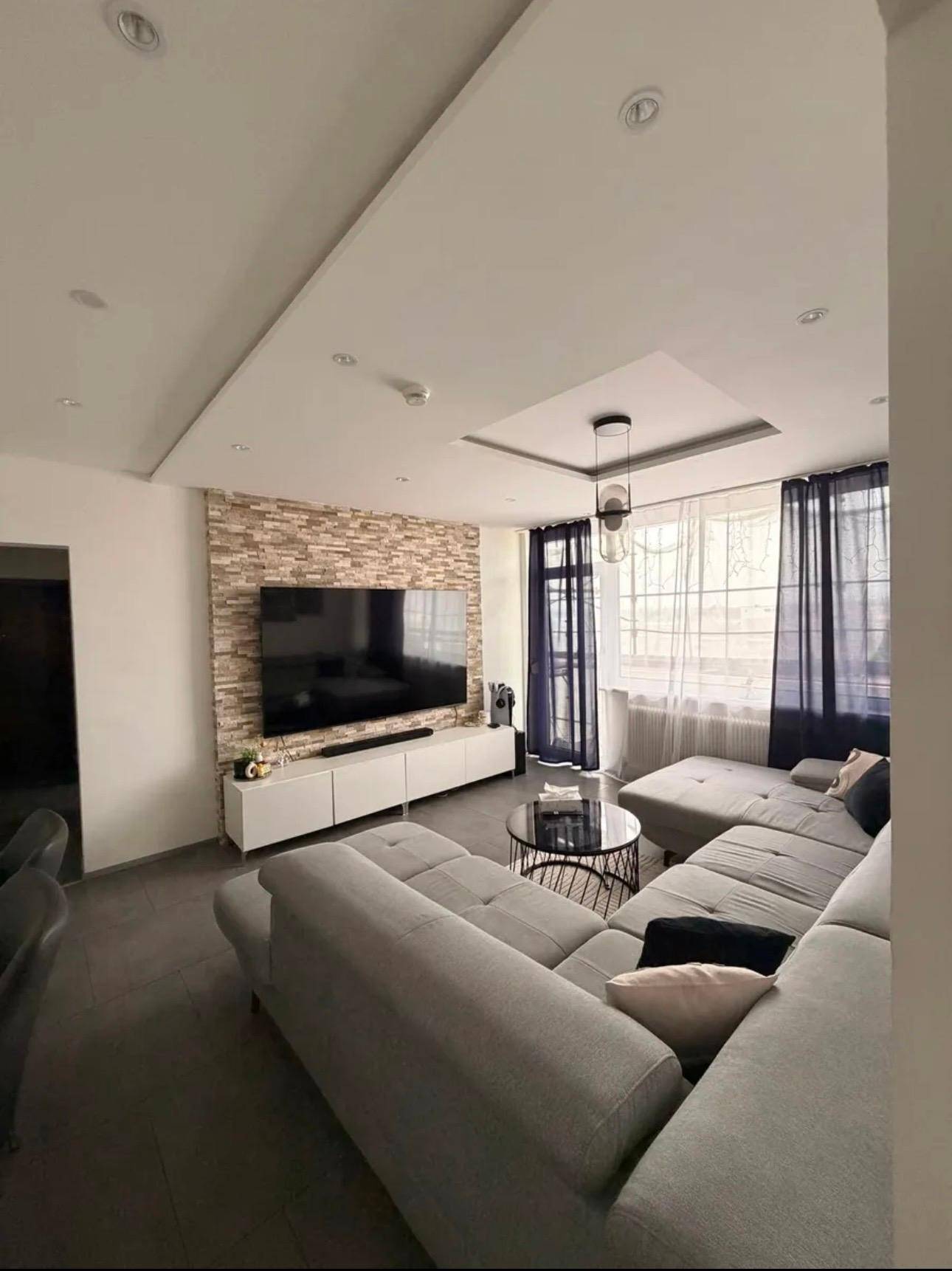 Geräumiges Wohnzimmer mit Ecksofa, Steinwand und viel Tageslicht durch große Fenster.