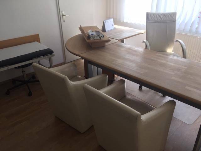 Büro- oder Behandlungszimmer mit Schreibtisch, Stühlen und Untersuchungsliege.