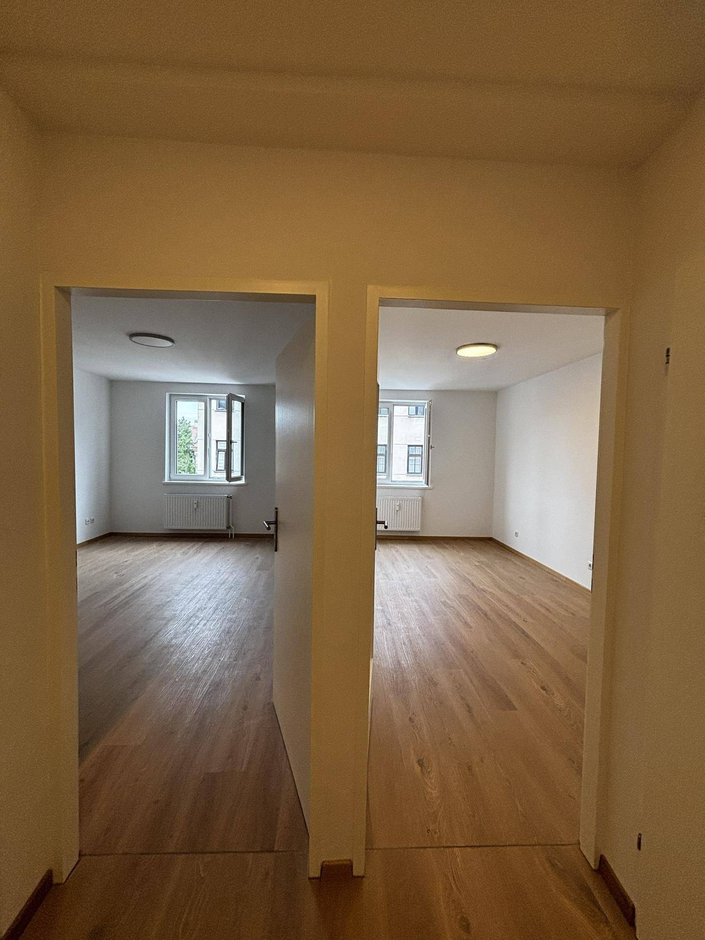 Flur mit Blick in zwei helle, leere Zimmer, beide mit Holzboden und Fenstern.