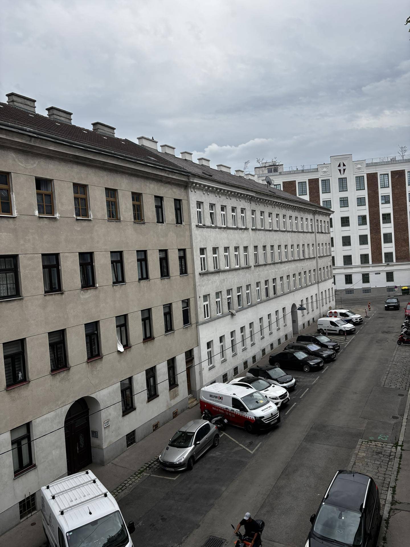 Blick aus dem Fenster auf eine belebte Straße mit parkenden Autos und umliegenden Gebäuden.