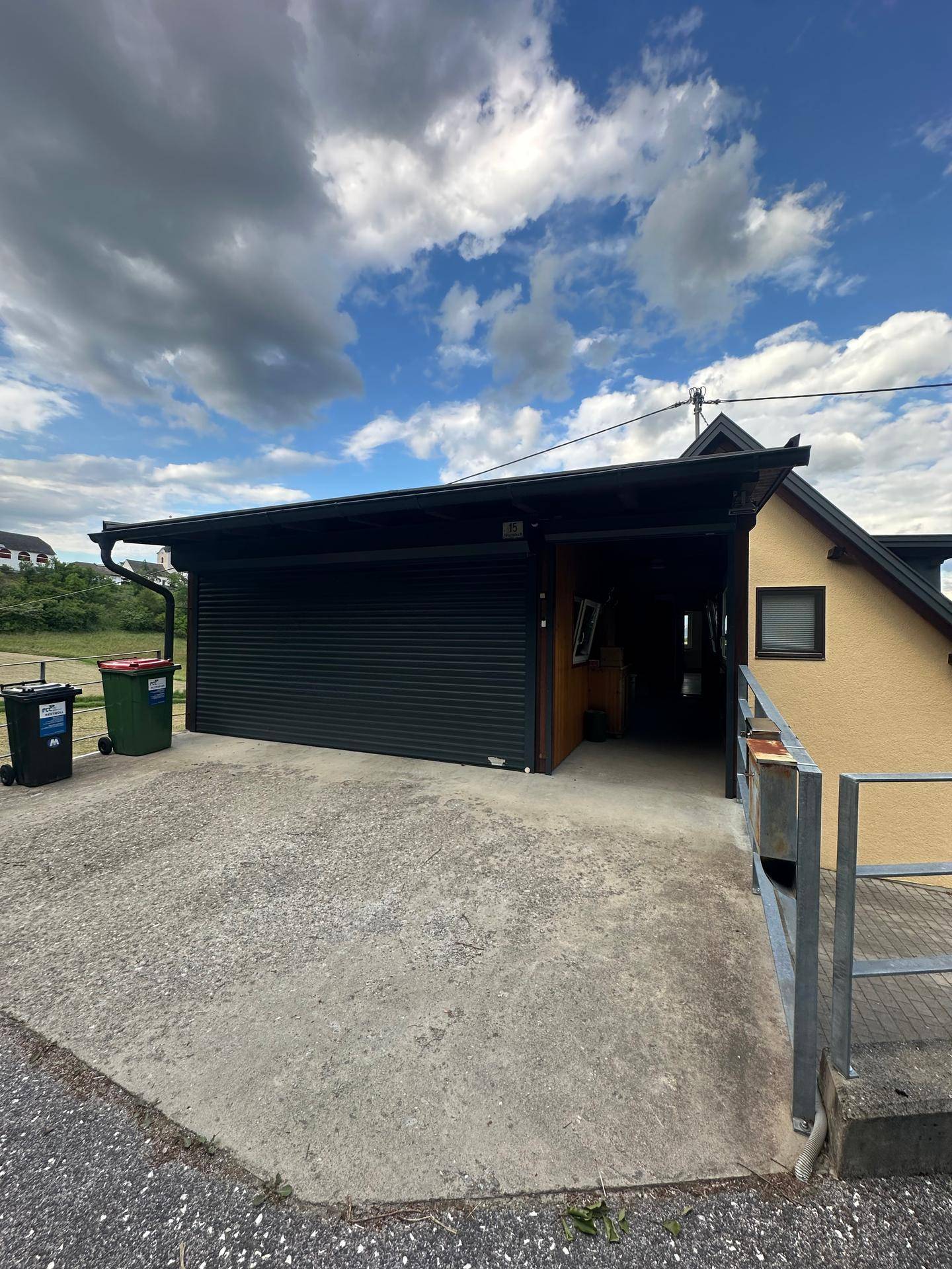 Überdachter Stellplatz oder Carport mit dunklem Rolltor und Mülltonnen.
