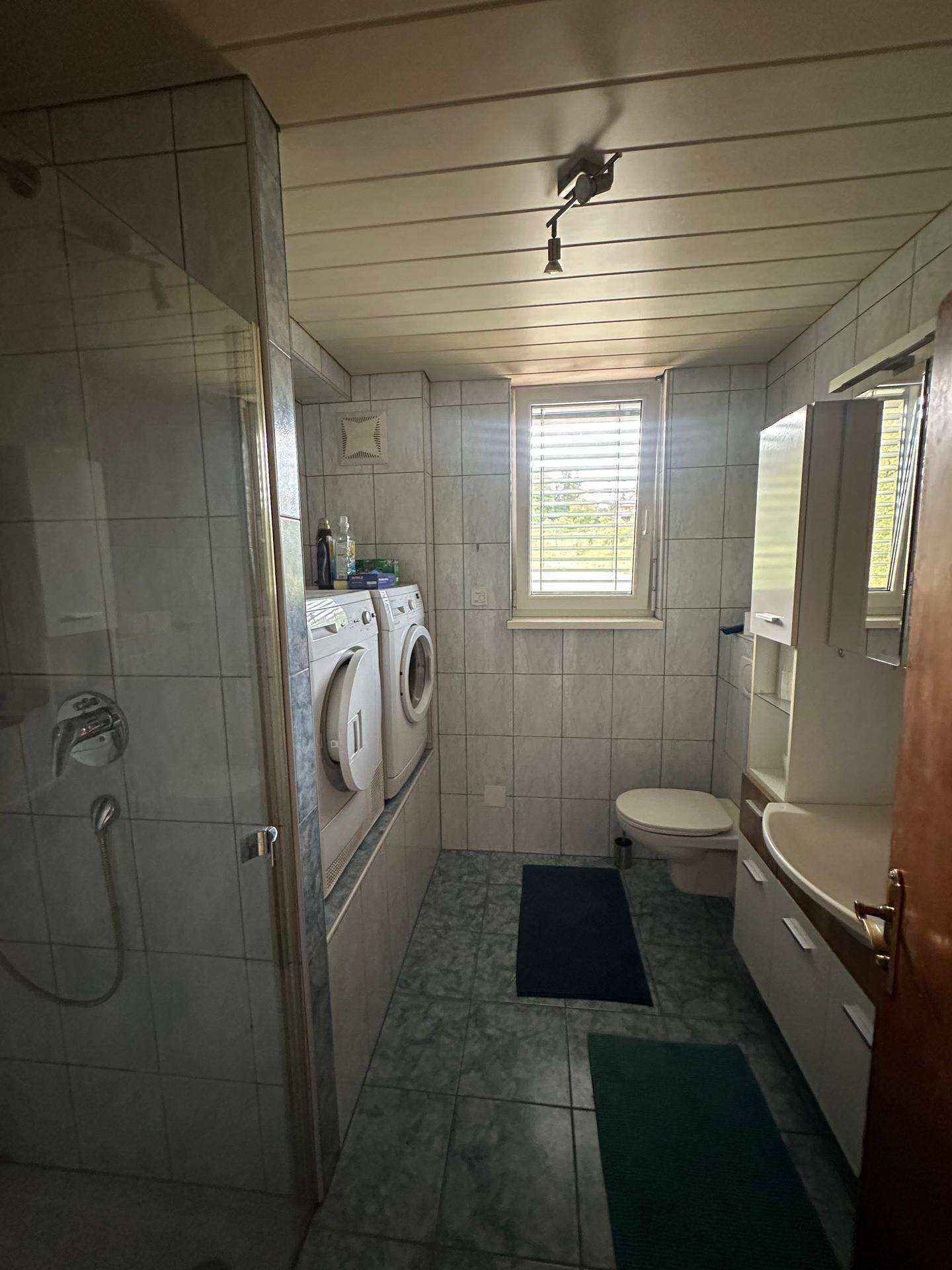 Badezimmer mit Dusche, WC, Waschbecken und zwei Waschmaschinen, gefliest.