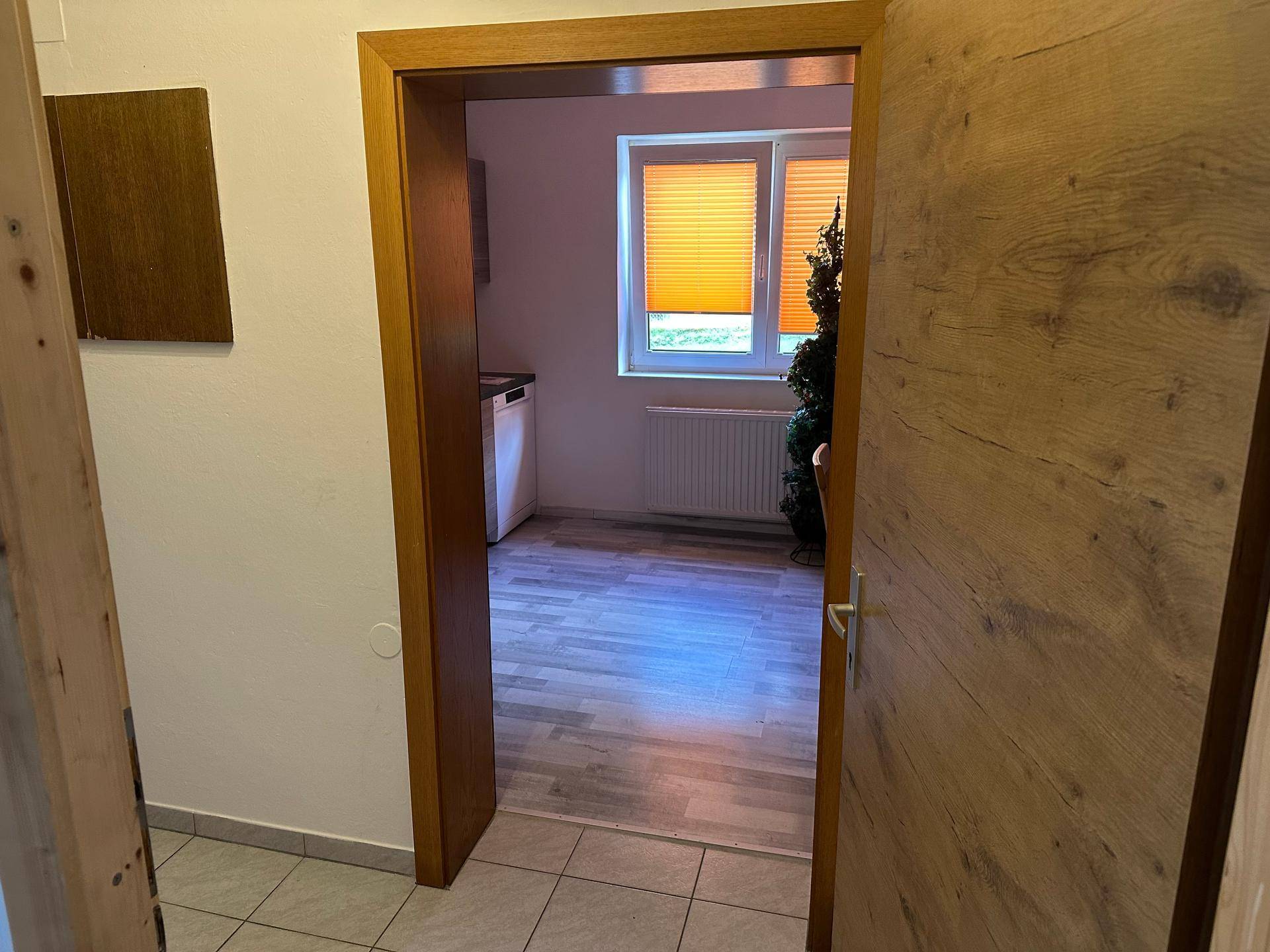 Blick durch eine Holztür in einen Raum mit Küchenzeile und hellem Boden.