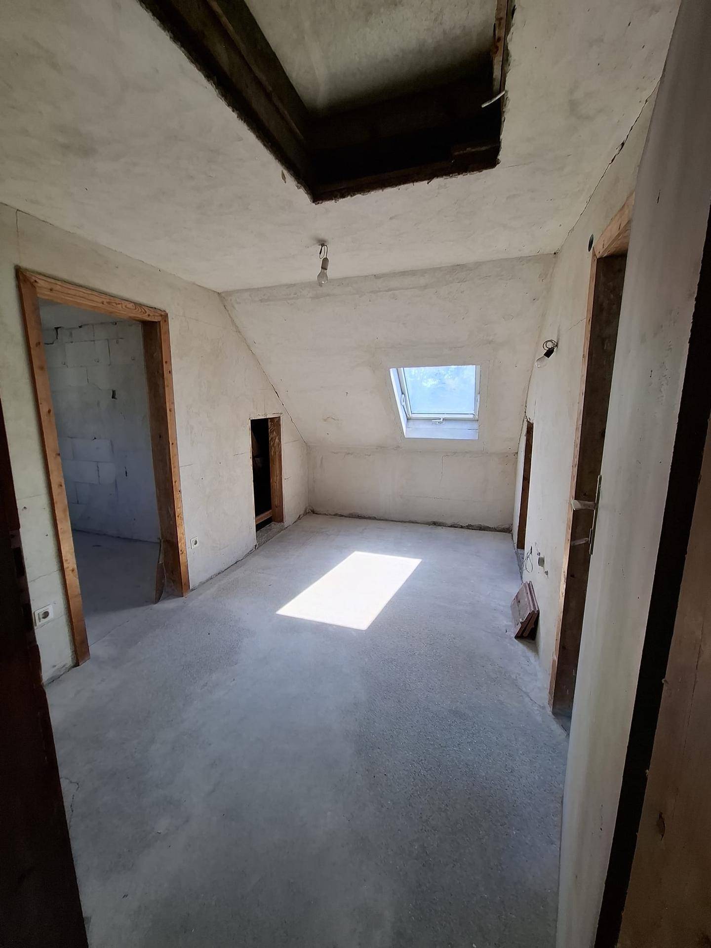 Dachgeschossraum im Rohbau mit Betonboden, schrägen Decken und einem Dachfenster.