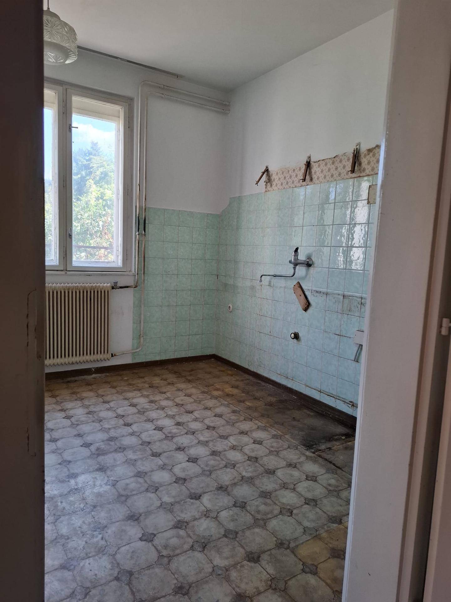 Leere Küche mit hellgrünen Wandfliesen, Fenster und Heizkörper, bereit für eine neue Einrichtung.