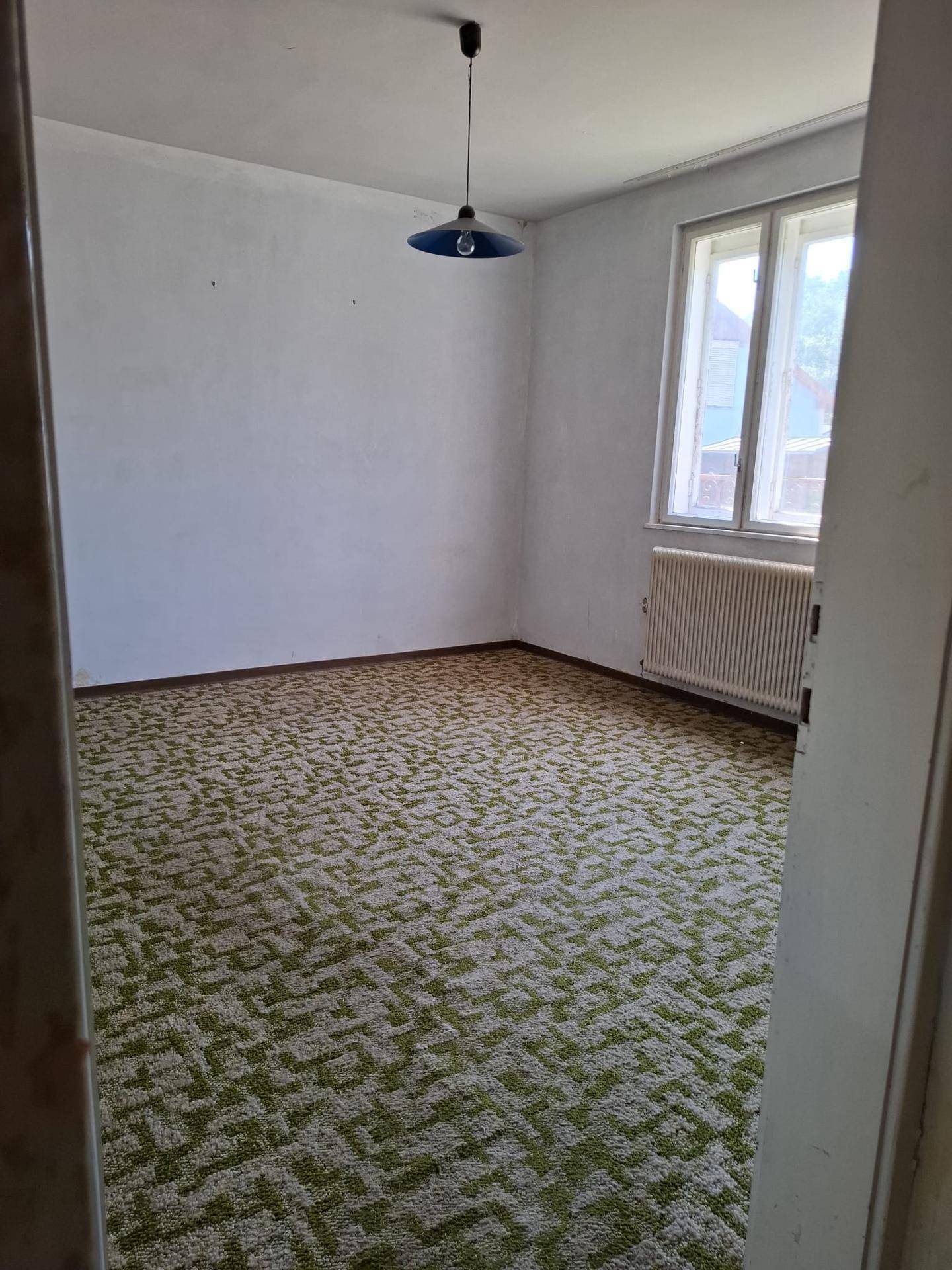 Schlafzimmer mit grün gemustertem Teppichboden, Fenster und Heizkörper, bereit zur Neugestaltung.