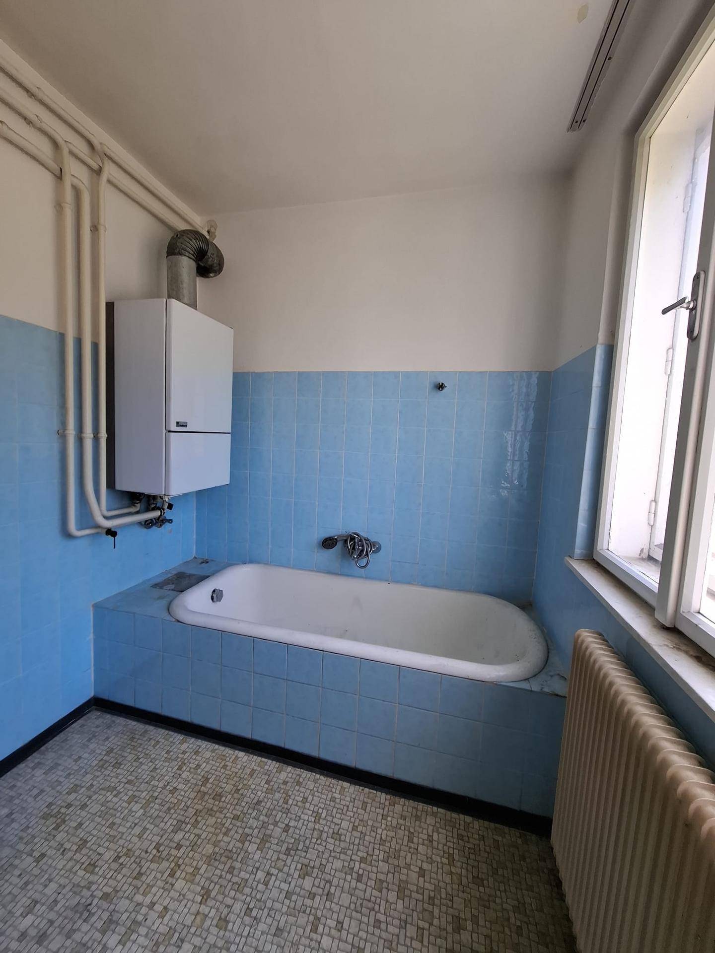 Badezimmer mit blauen Wandfliesen, Badewanne, Fenster und einem an der Wand montierten Warmwasserbereiter.