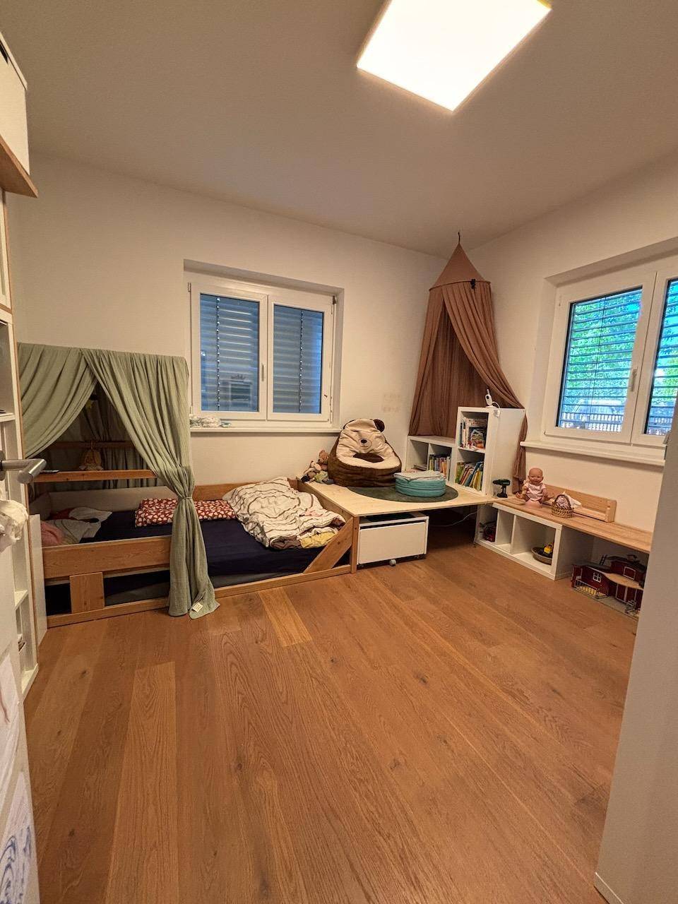 Kinderzimmer mit zwei Betten, Holzfußboden und hellen Fenstern mit Jalousien.