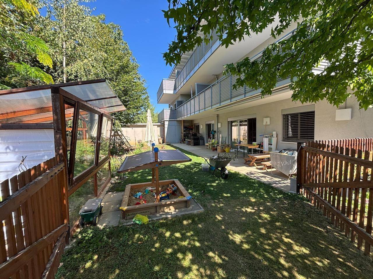 Weitläufiger Garten mit Gewächshaus, Sandkasten und Terrasse am Gebäude.
