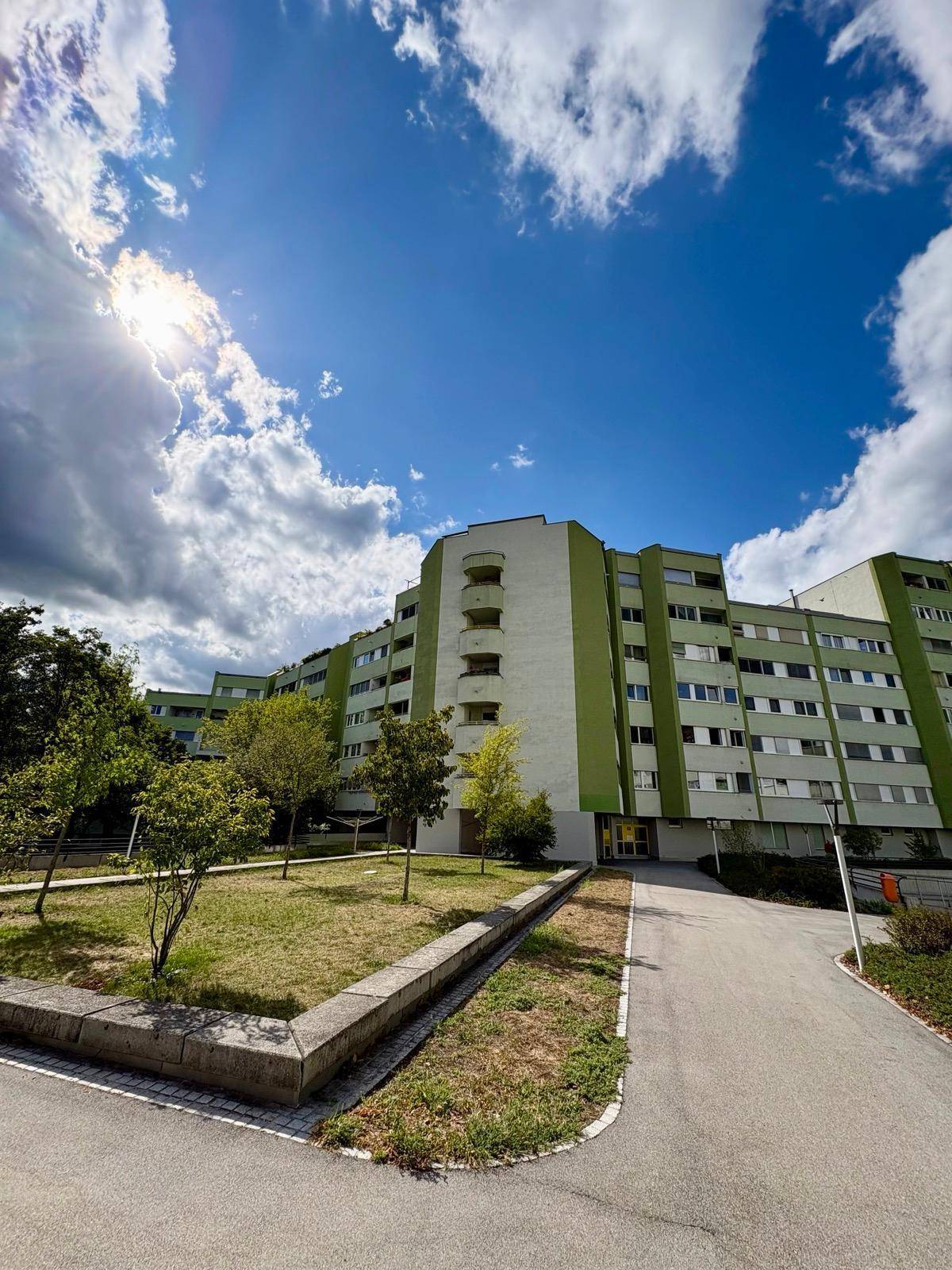 Mehrstöckiges Wohngebäude mit grüner Fassade und Bäumen unter blauem Himmel.