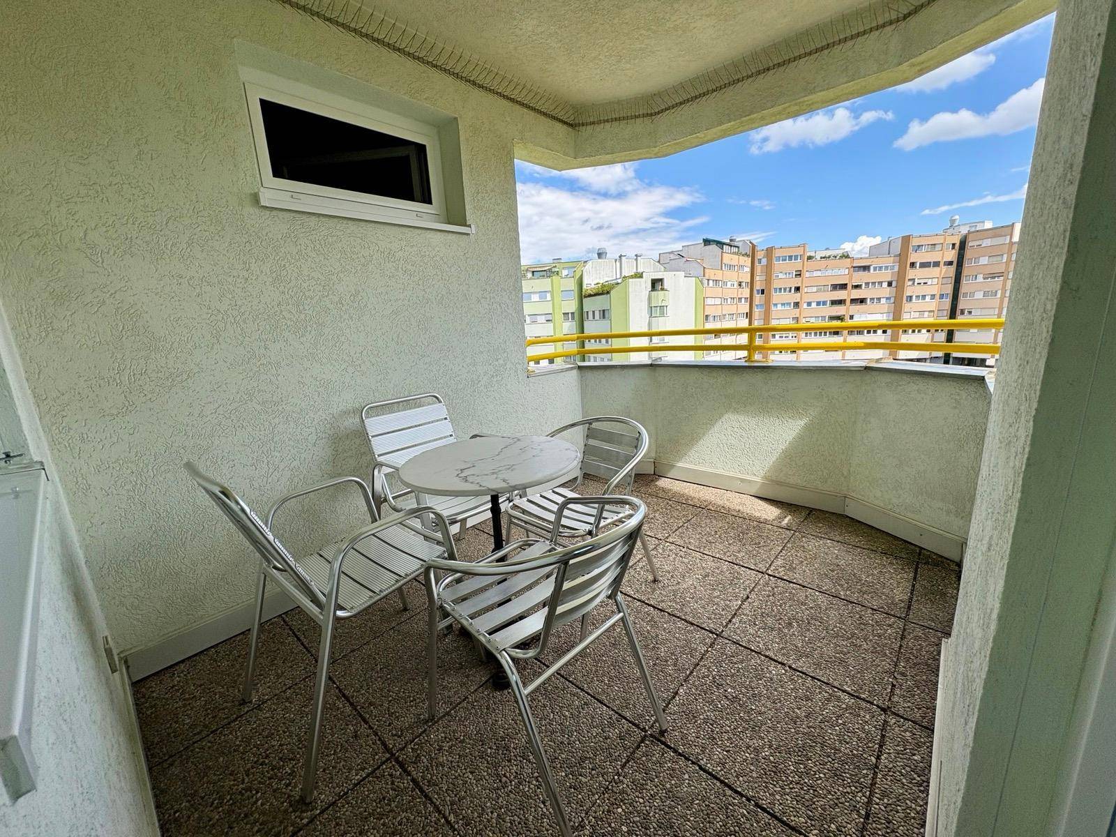 Kleiner Balkon mit Fliesenboden und Sitzgelegenheiten, umgeben von Stadtgebäuden.