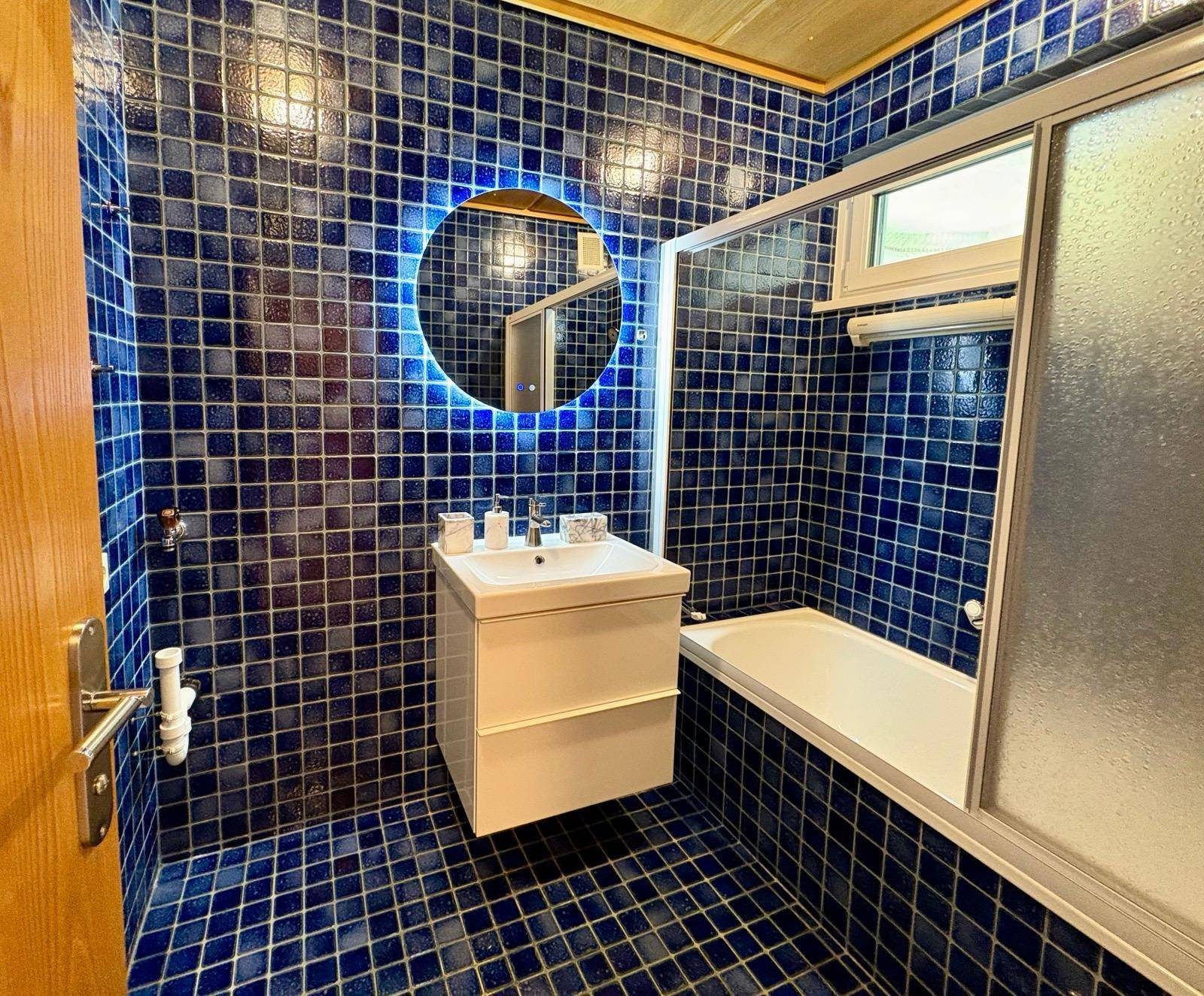 Badezimmer mit blauen Fliesen, Badewanne mit Duschabtrennung und beleuchtetem Spiegel.
