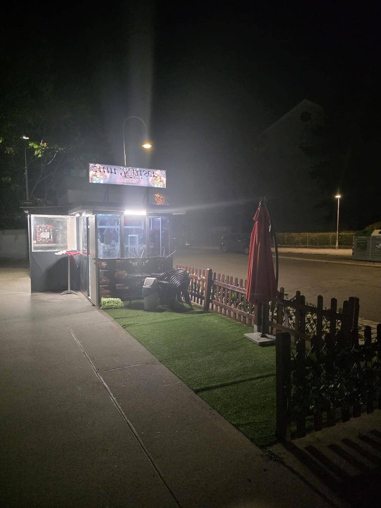 Nächtliche Ansicht eines beleuchteten Imbissstandes mit rotem Sonnenschirm und kleiner Grünfläche.