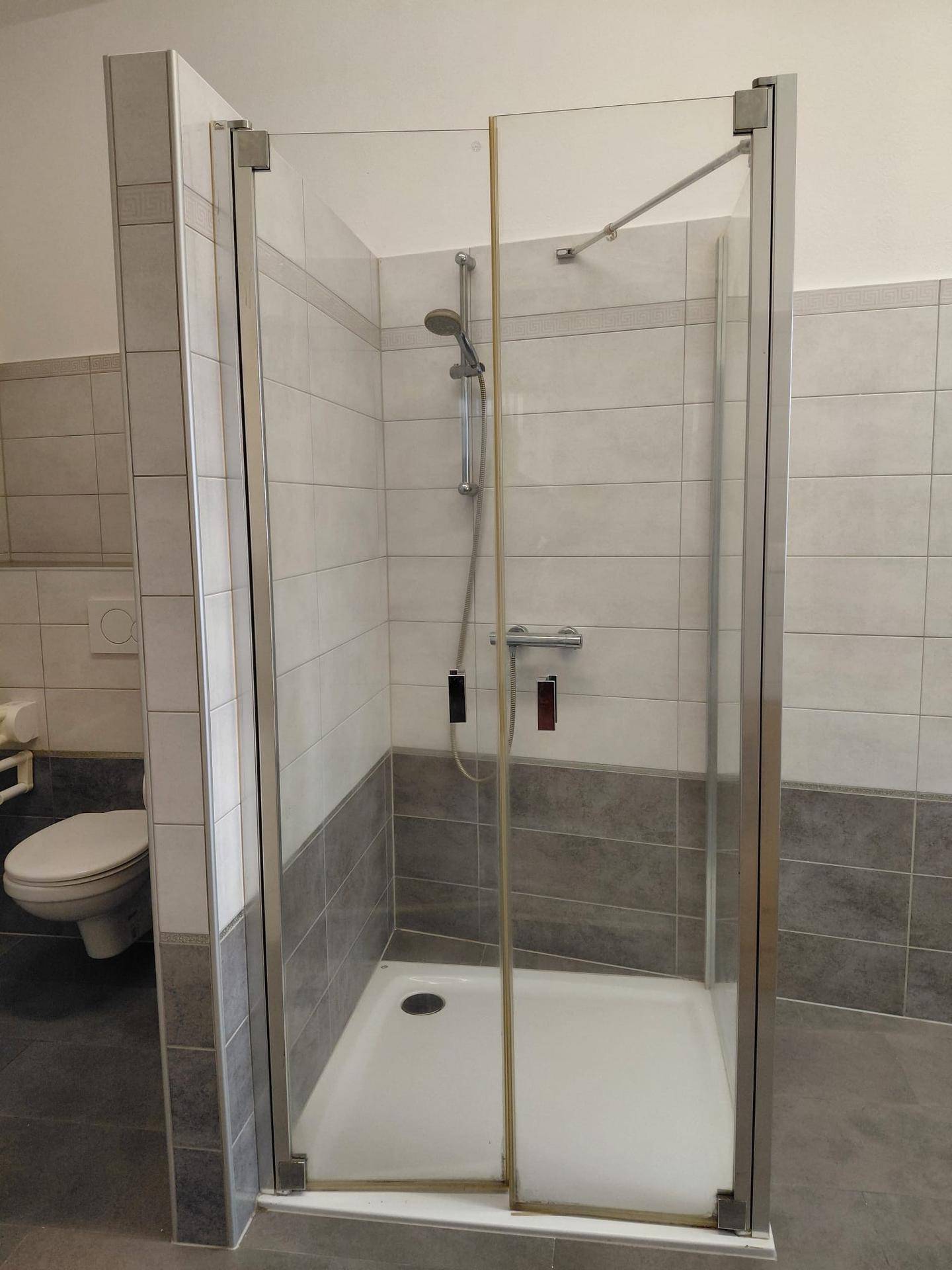Duschbereich im Badezimmer mit Glasabtrennung und Wandfliesen in zwei Grautönen.