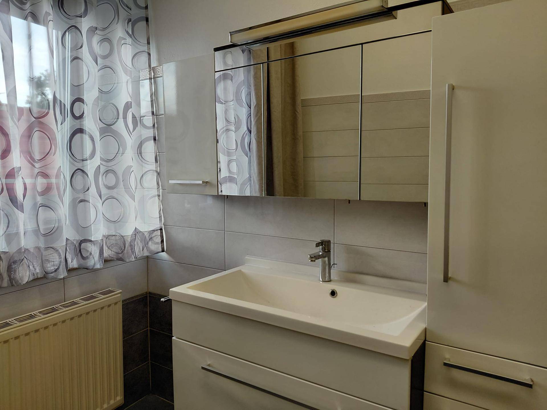 Badezimmer mit modernem Waschtisch, Spiegelschrank und Heizkörper, gefliest in Grau und Weiß.