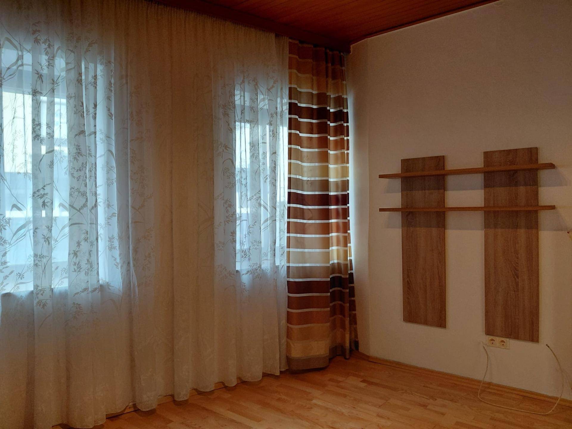 Helles Zimmer mit Holzdecke, Parkettboden und zwei Fenstern mit Vorhängen.