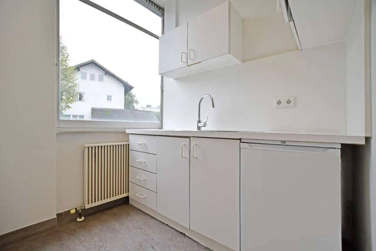 Kleine Küchenzeile mit weißen Schränken, Spüle und Kühlschrank unter einem Fenster mit Heizkörper.