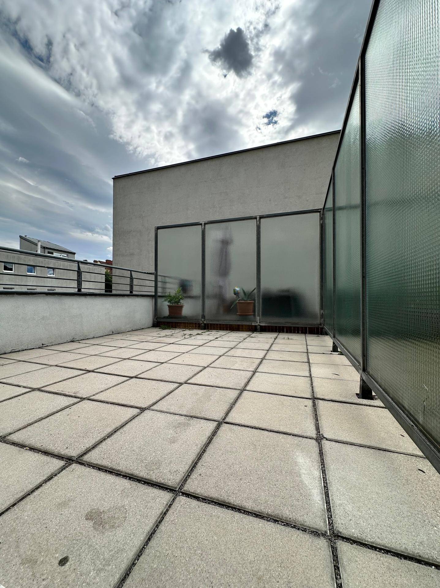 Geräumige Dachterrasse mit Fliesenboden und Milchglas-Trennwänden unter bewölktem Himmel.