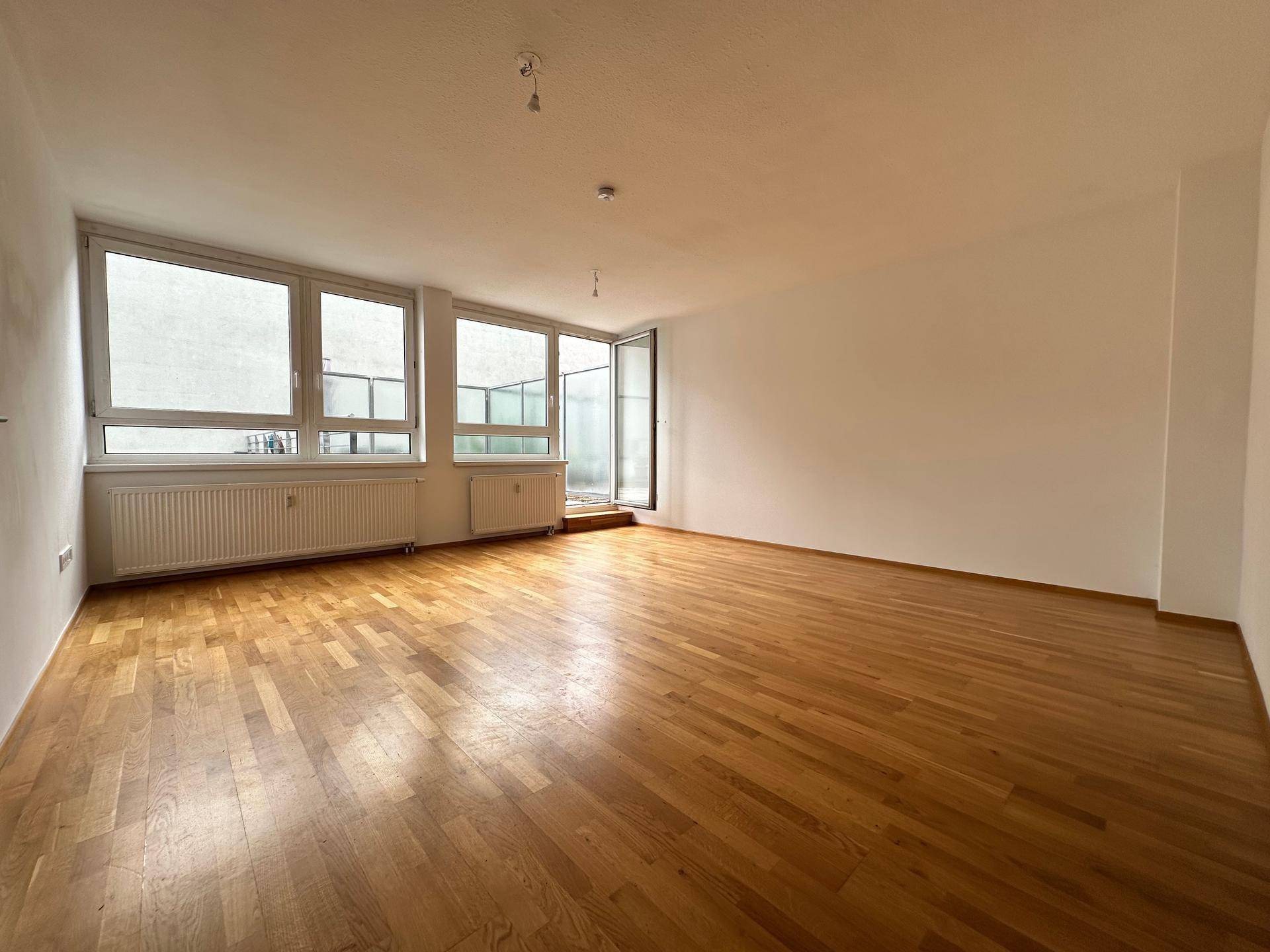 Leeres Wohnzimmer mit Parkettboden, großen Fenstern und Zugang zu einem Außenbereich.