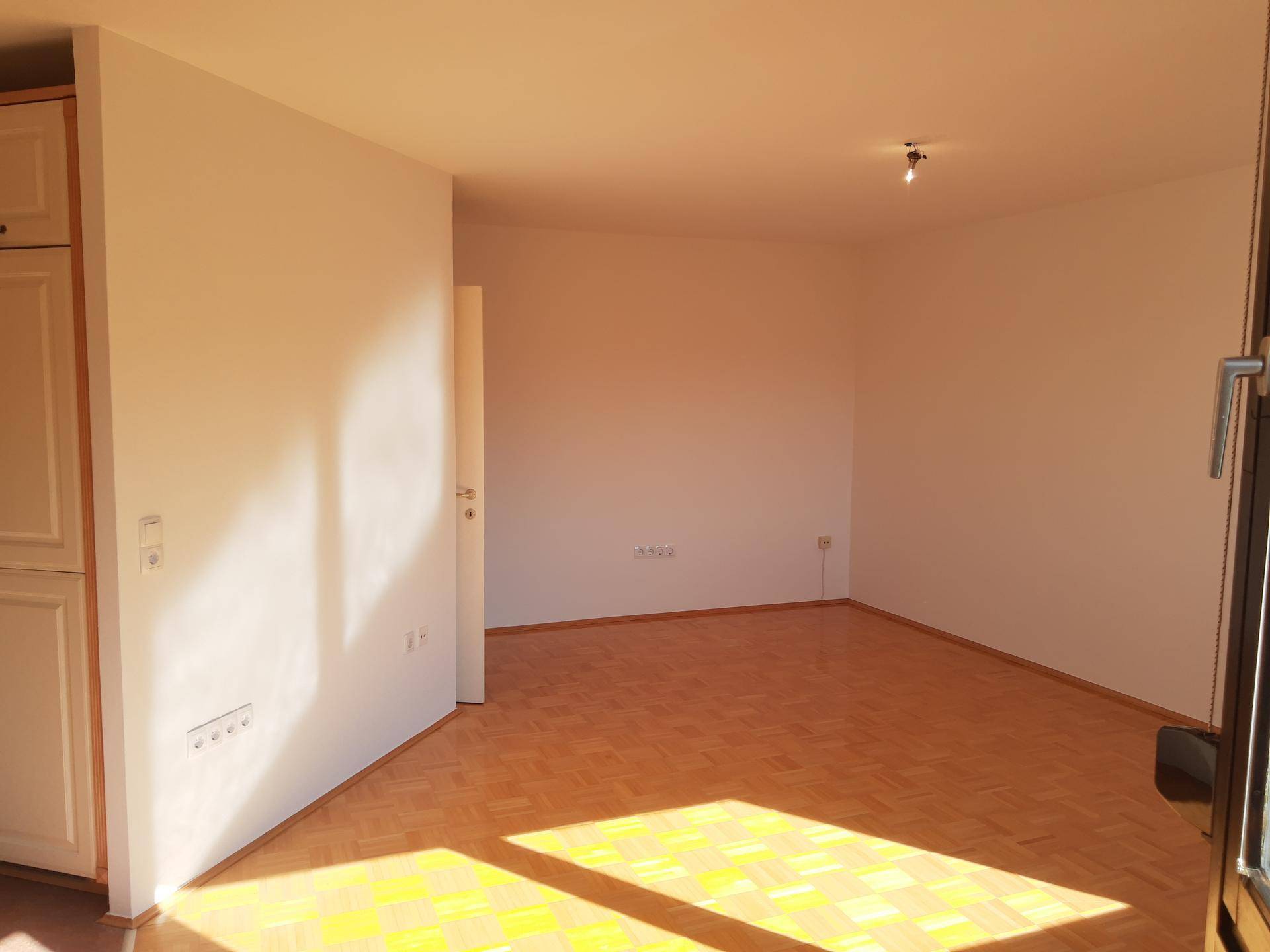 Heller, leerer Raum mit Parkettboden und weiß gestrichenen Wänden, durchflutet von Sonnenlicht.