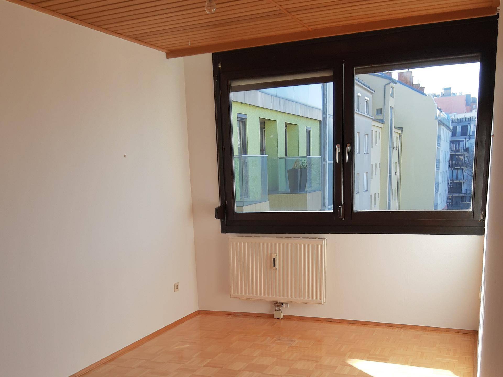 Helles Schlafzimmer mit Parkettboden, Holzdecke und Fensterblick auf die umliegenden Gebäude.
