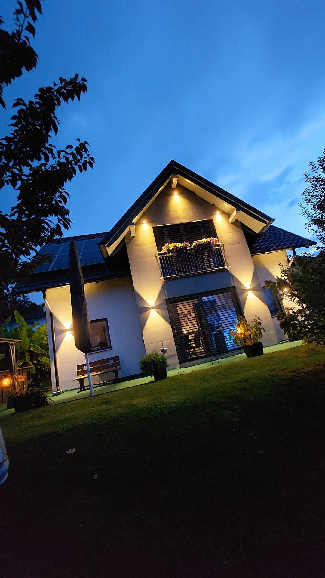Das Haus bei Nacht mit stimmungsvoller Außenbeleuchtung und beleuchtetem Balkon.