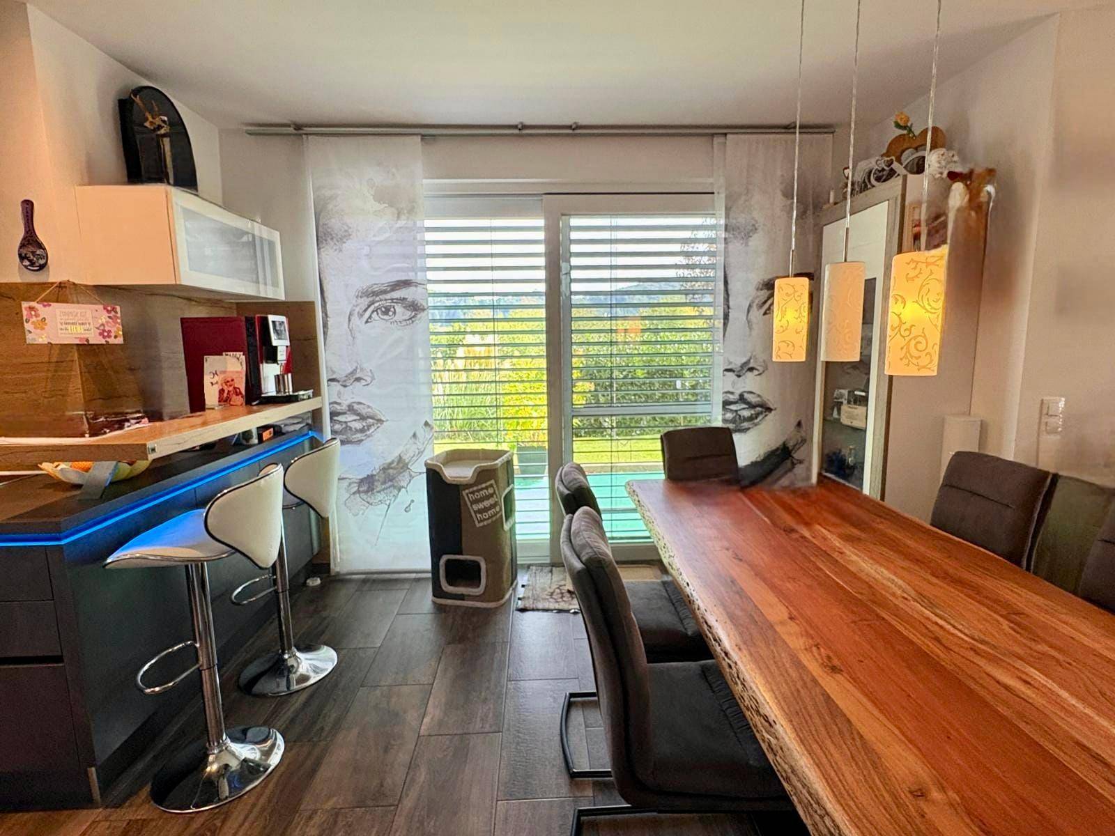 Moderne Küche mit Bartheke, Holztisch und Blick ins Grüne durch große Fenster.