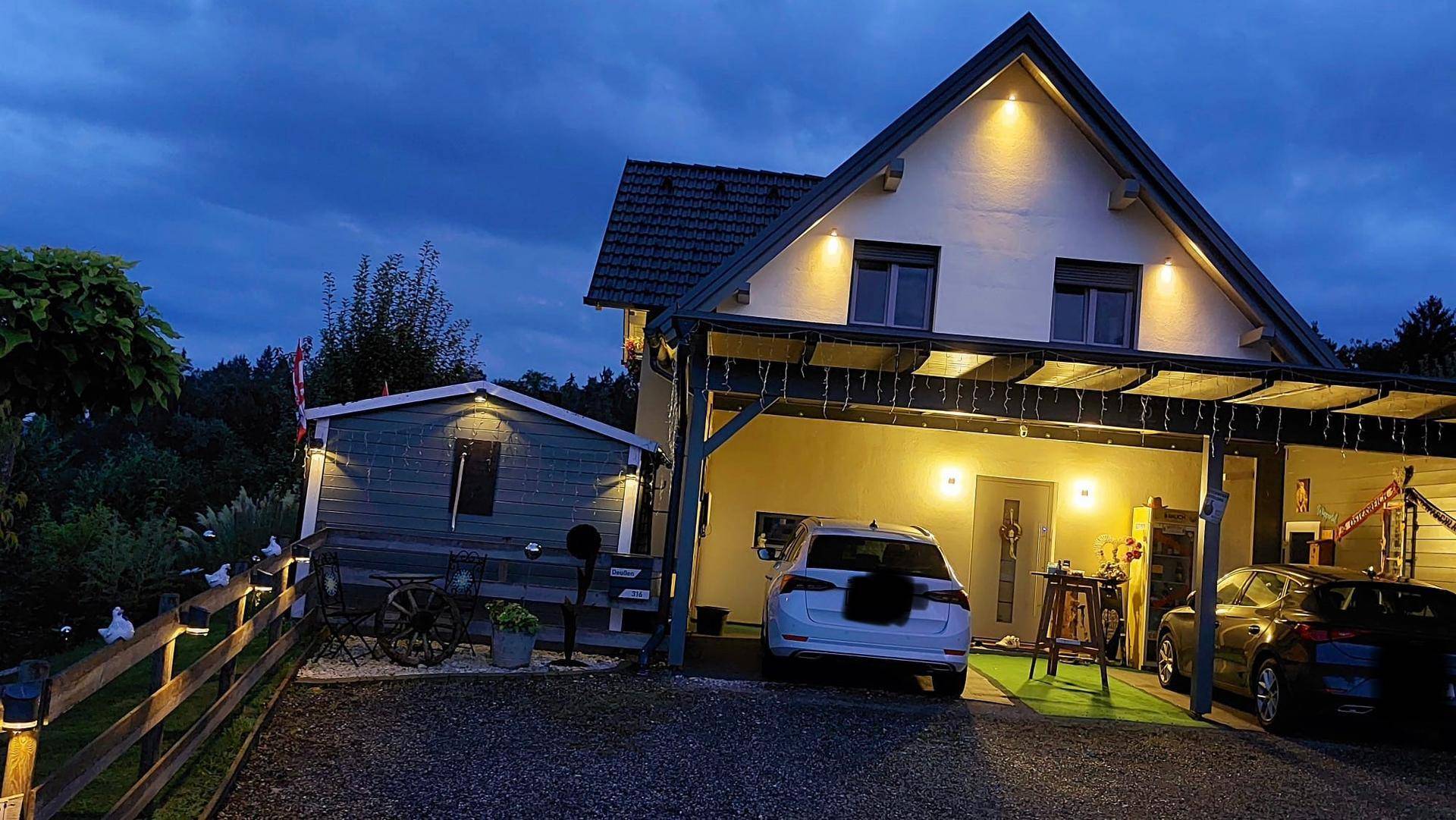 Hausfront bei Nacht mit beleuchtetem Carport und geparkten Fahrzeugen.