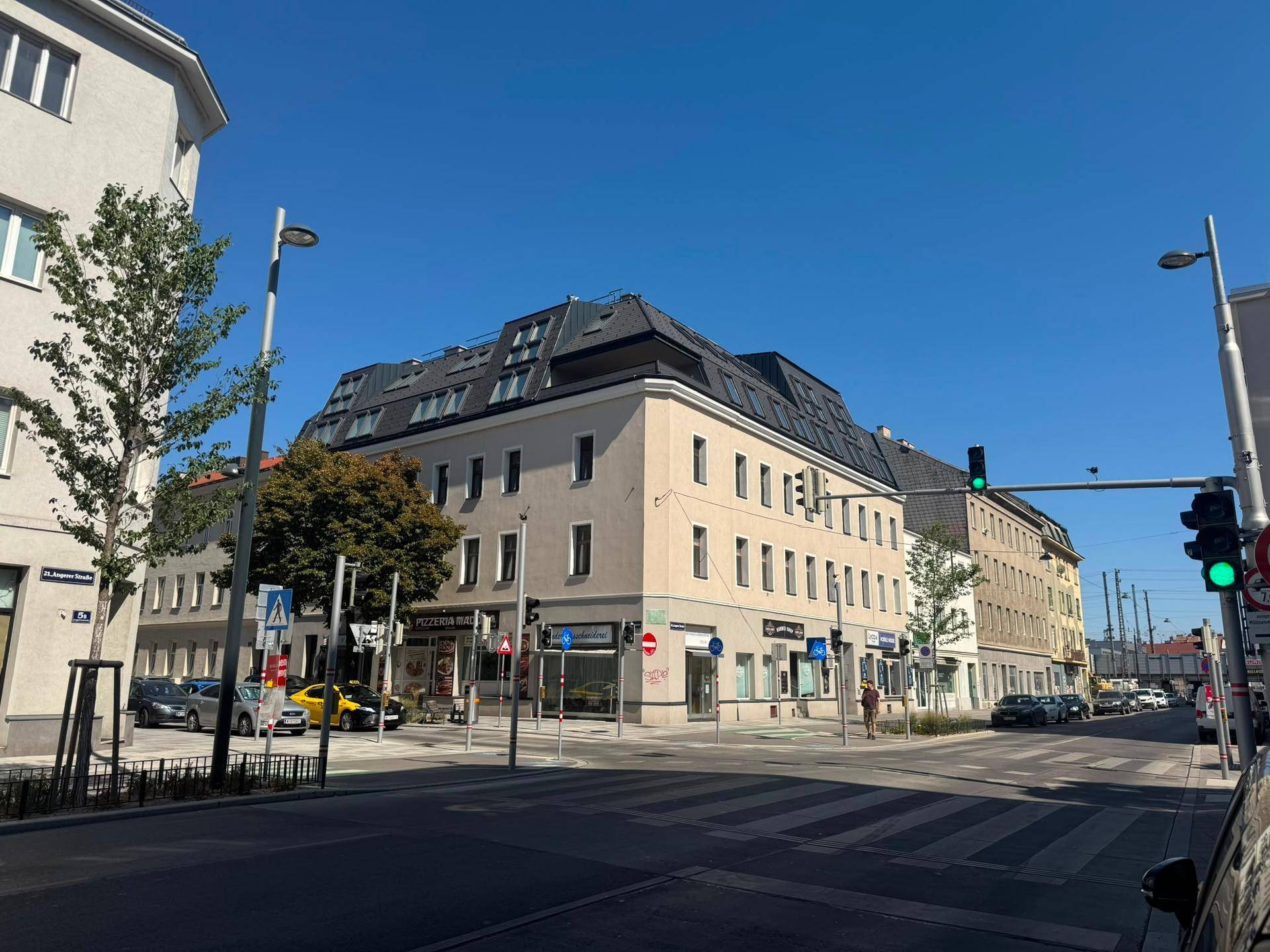 Straßenecke mit mehrstöckigen Gebäuden, Geschäften im Erdgeschoss und einem Mansarddach unter blauem Himmel.