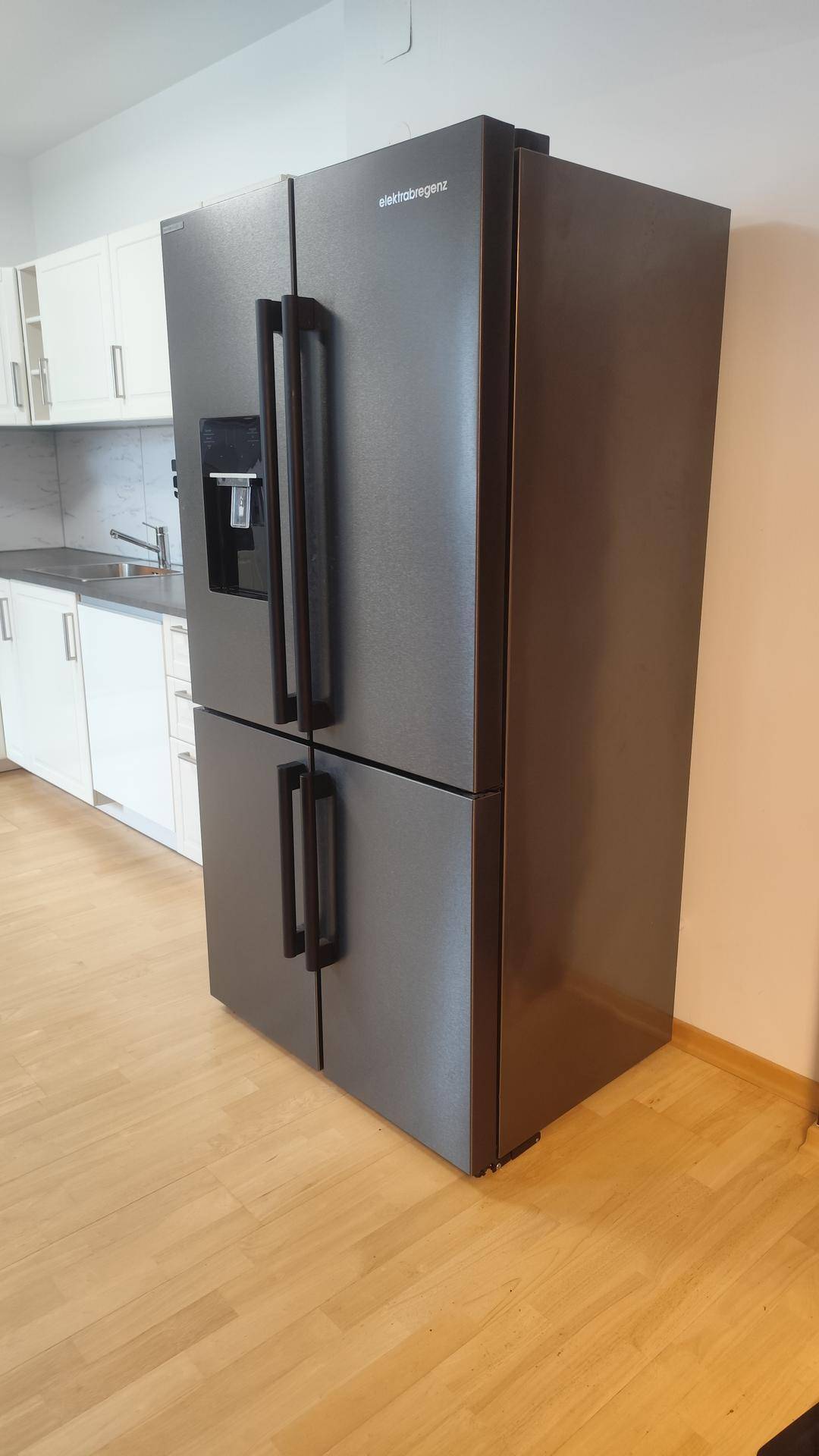 Großer, moderner schwarzer Kühlschrank mit Wasserspender auf hellem Holzfußboden.