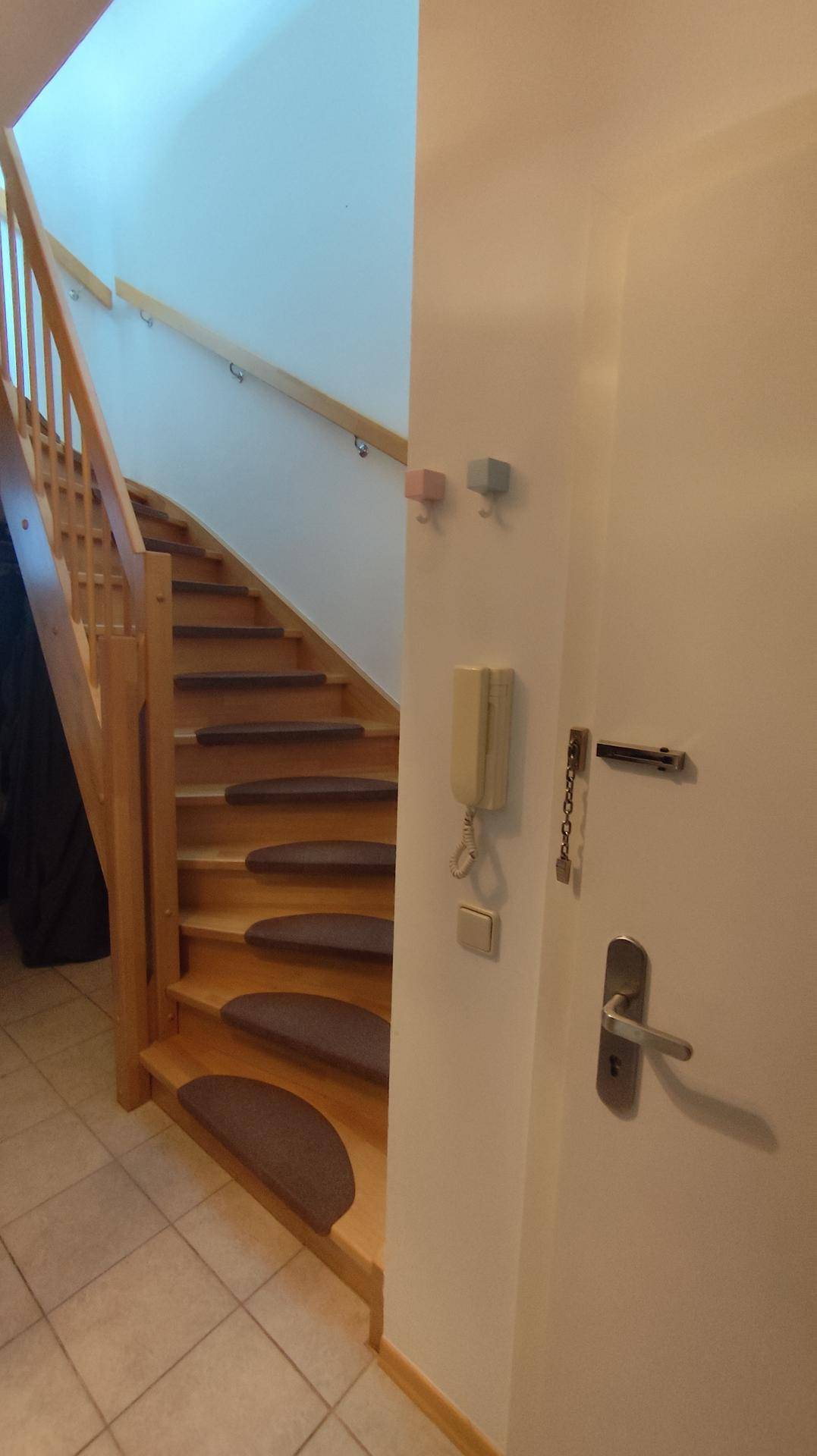 Eingangsbereich mit Fliesenboden, Holztreppe und Gegensprechanlage, die den Zugang zur Wohnung regelt.