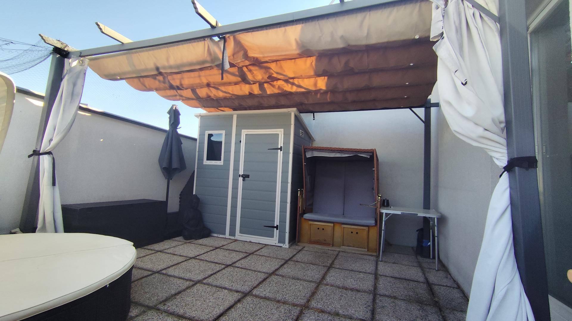 Geräumige Dachterrasse mit Pergola, Gartenhaus, Strandkorb und Whirlpool unter blauem Himmel.