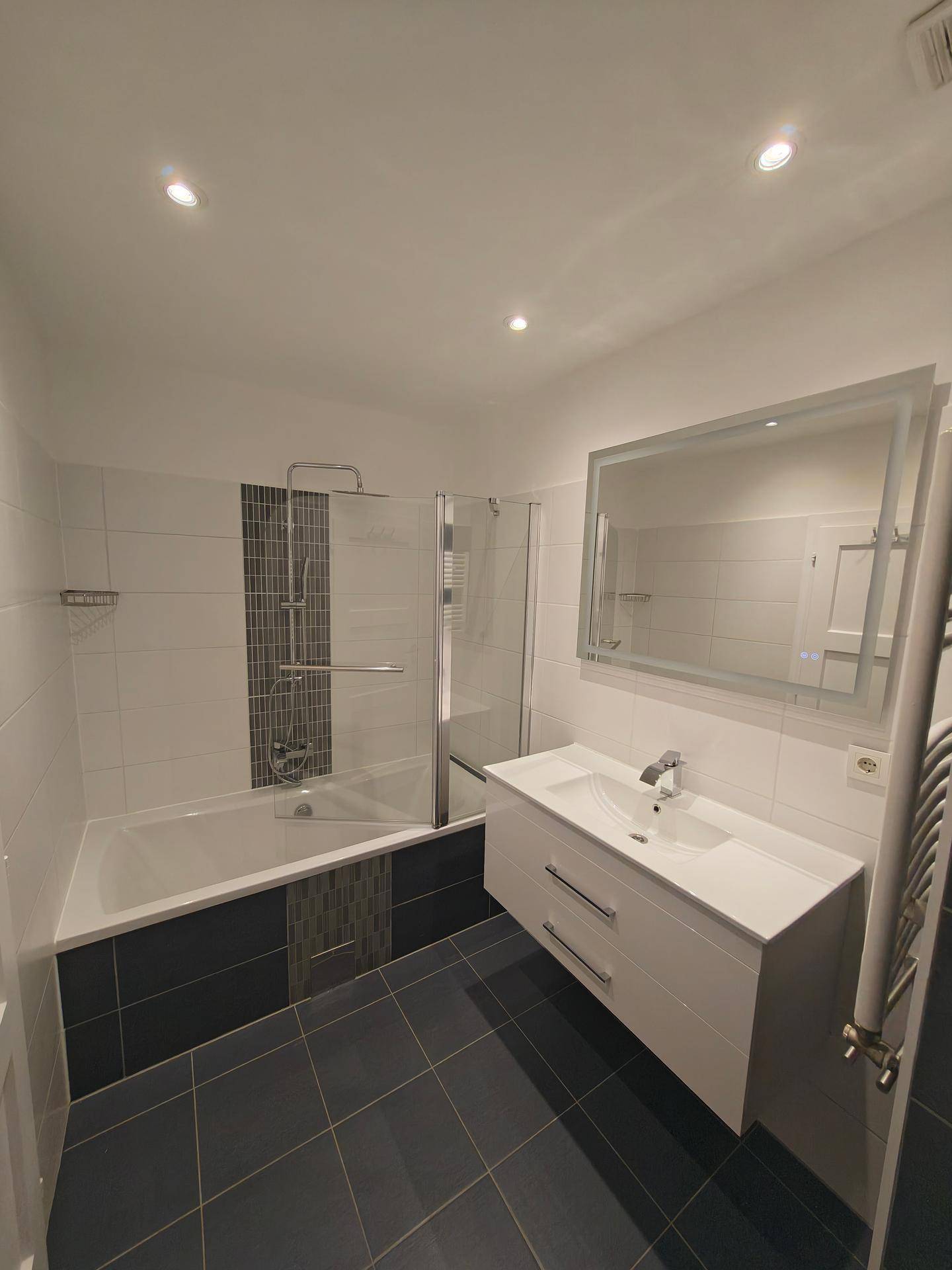 Modernes Badezimmer mit Badewanne, Duschwand, Einzelwaschbecken und beleuchtetem Spiegel.