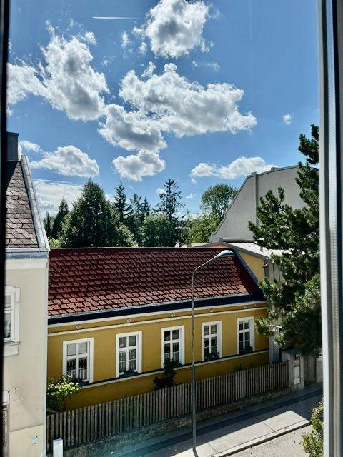 Blick aus dem Fenster auf ein charmantes gelbes Gebäude mit rotem Dach und Bäumen.