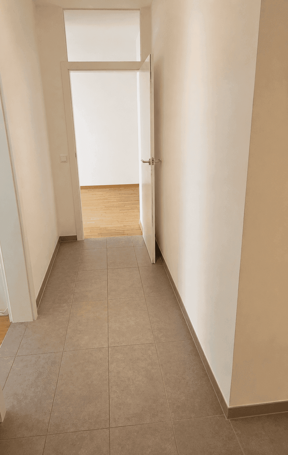 Heller Flur mit Fliesenboden und offener Tür, die zu einem Zimmer mit Parkett führt.