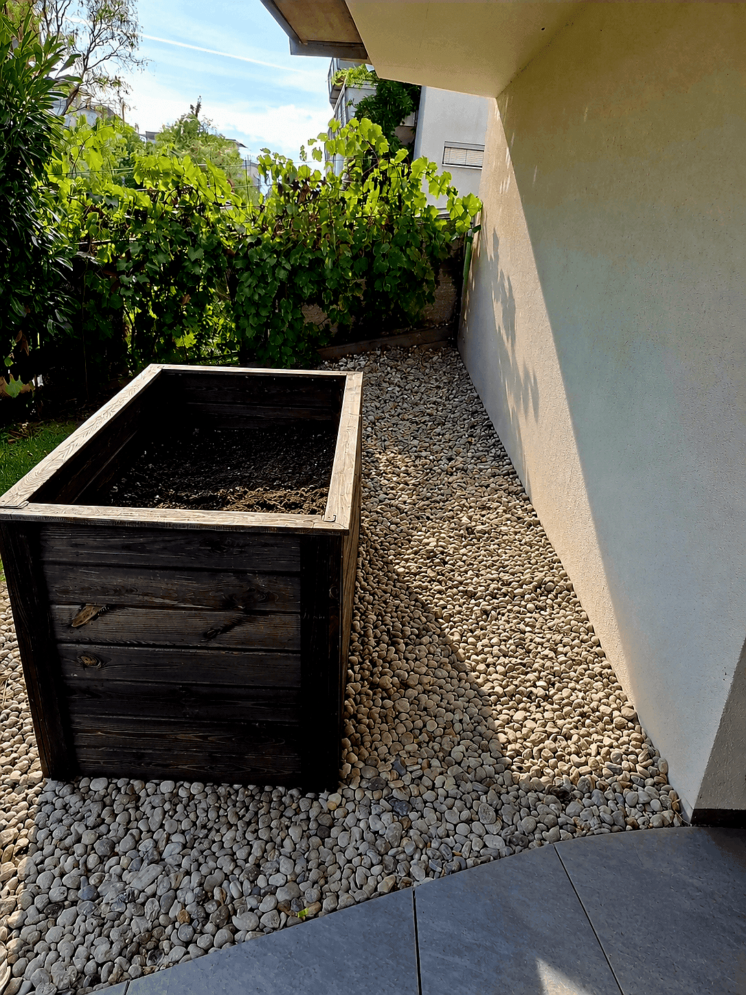 Gartenbereich mit Zierkies, einem hölzernen Pflanzkasten und einer hellen Hauswand im Sonnenlicht.