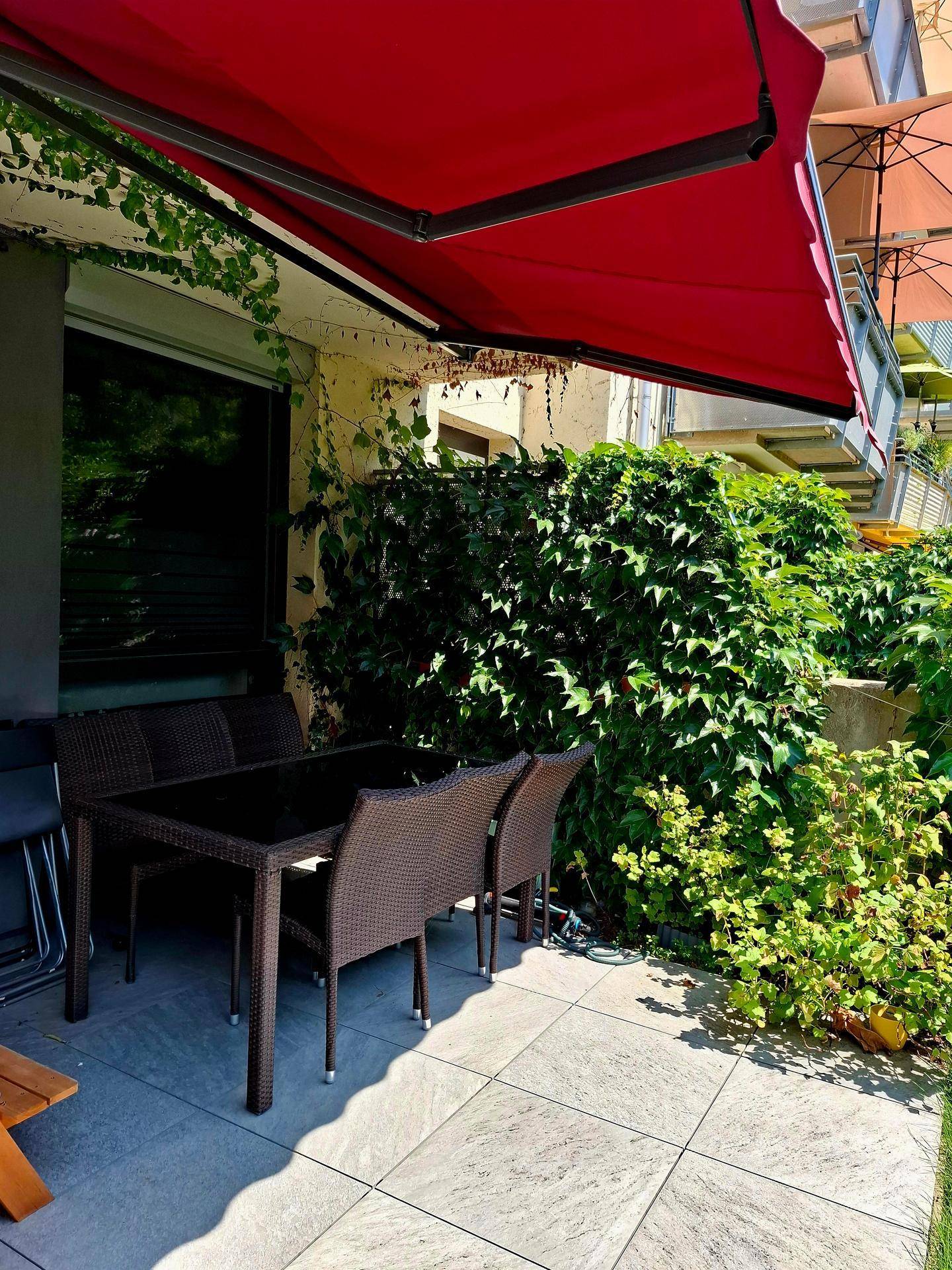 Gemütliche Terrasse mit Gartenmöbeln und roter Markise, umgeben von üppigem Grün.