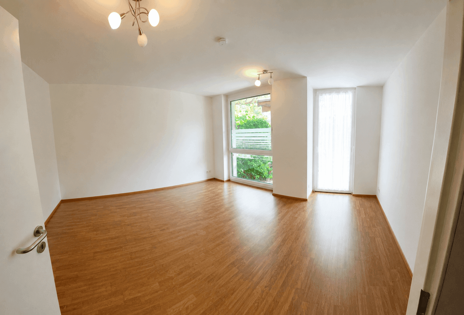 Helles, leeres Zimmer mit Holzboden und großen Fenstern, ideal für Schlaf- oder Wohnbereich.