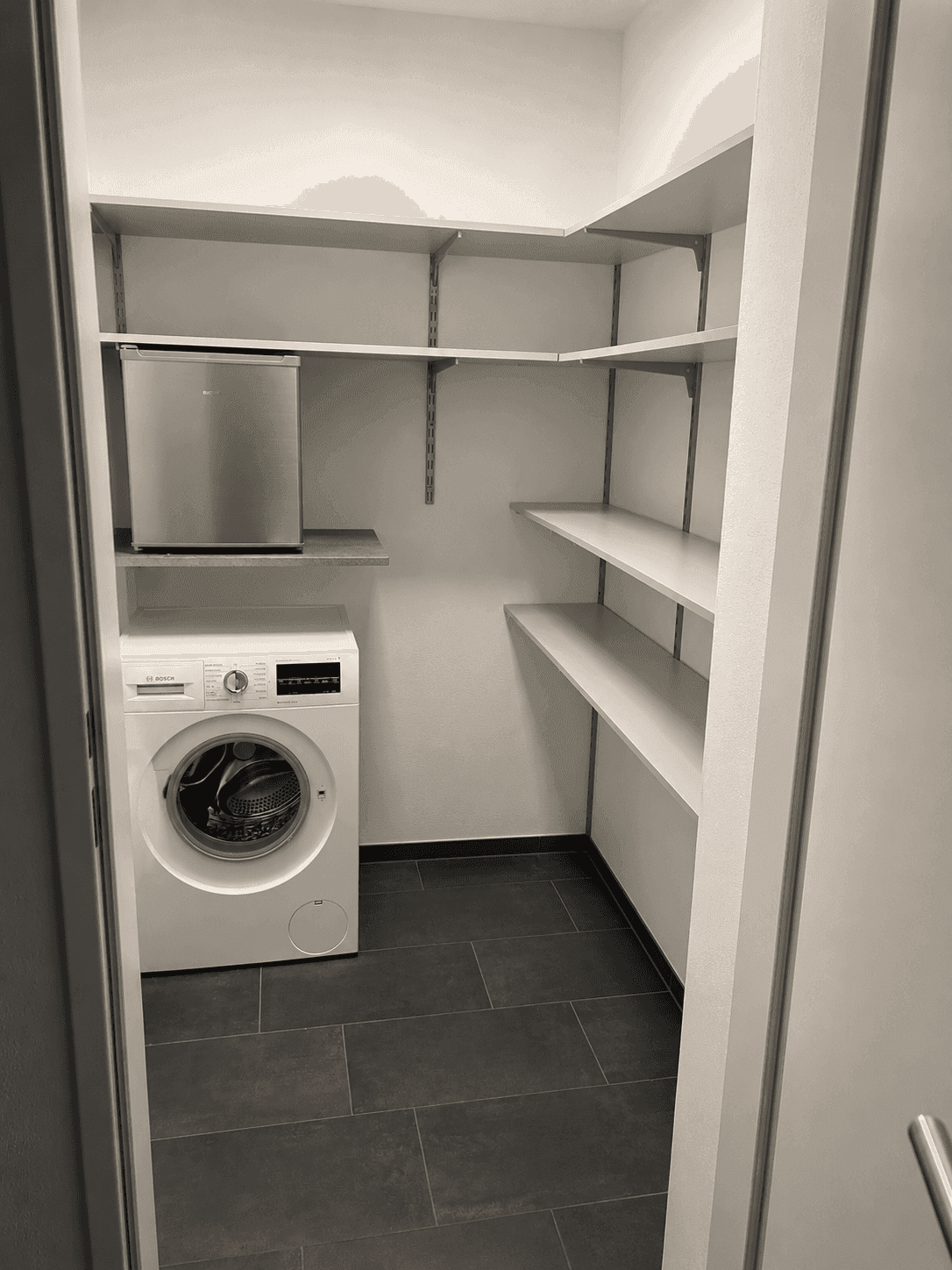 Praktischer Abstellraum mit Waschmaschine, Regalen und kleinem Kühlschrank.