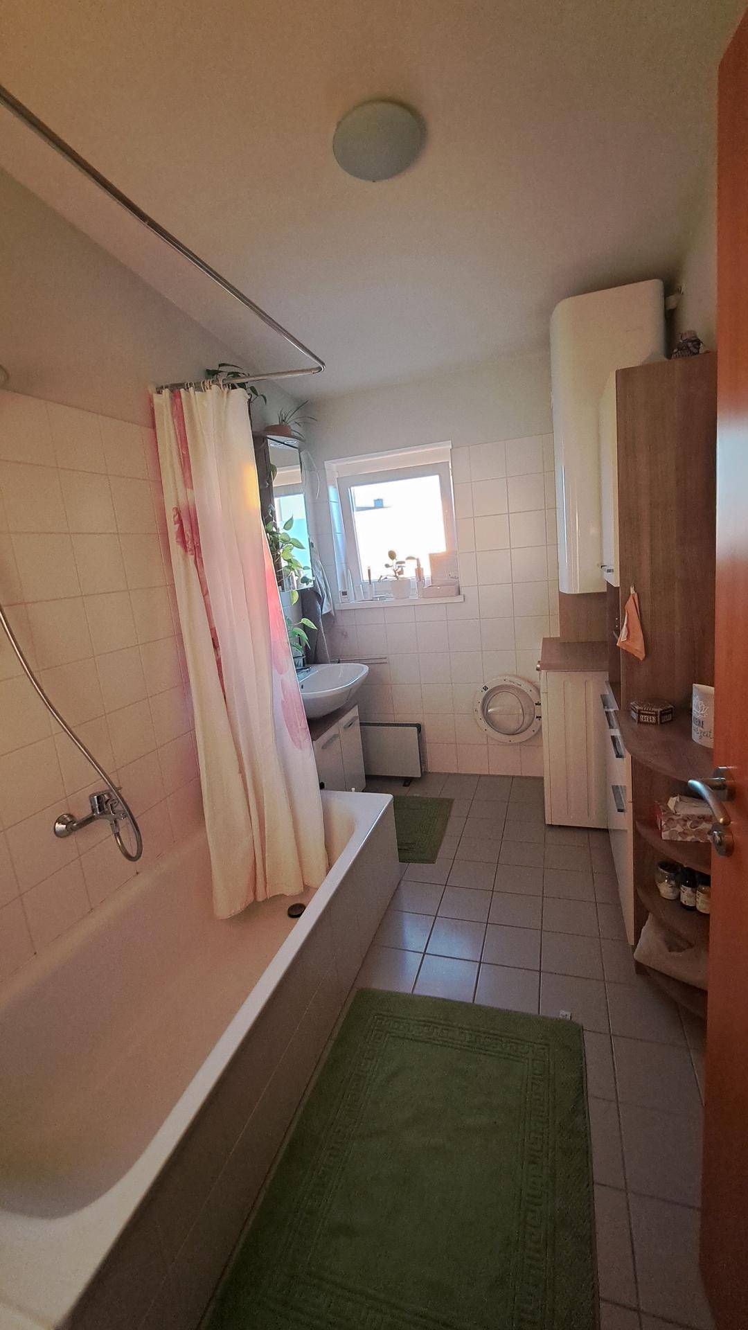 Gefliestes Badezimmer mit Badewanne, Duschvorhang, Waschbecken und Fenster für Tageslicht.