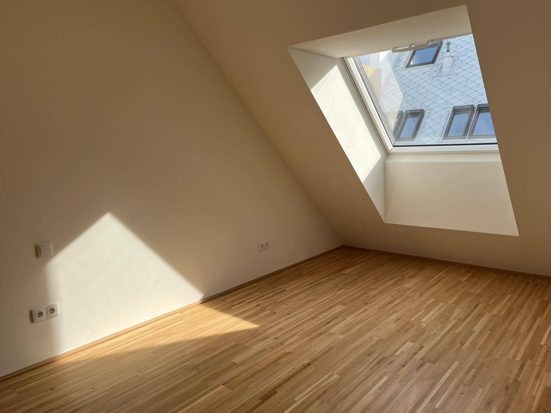 Helles Zimmer mit Holzboden, Dachschräge und großem Dachfenster, durchflutet von Sonnenlicht.