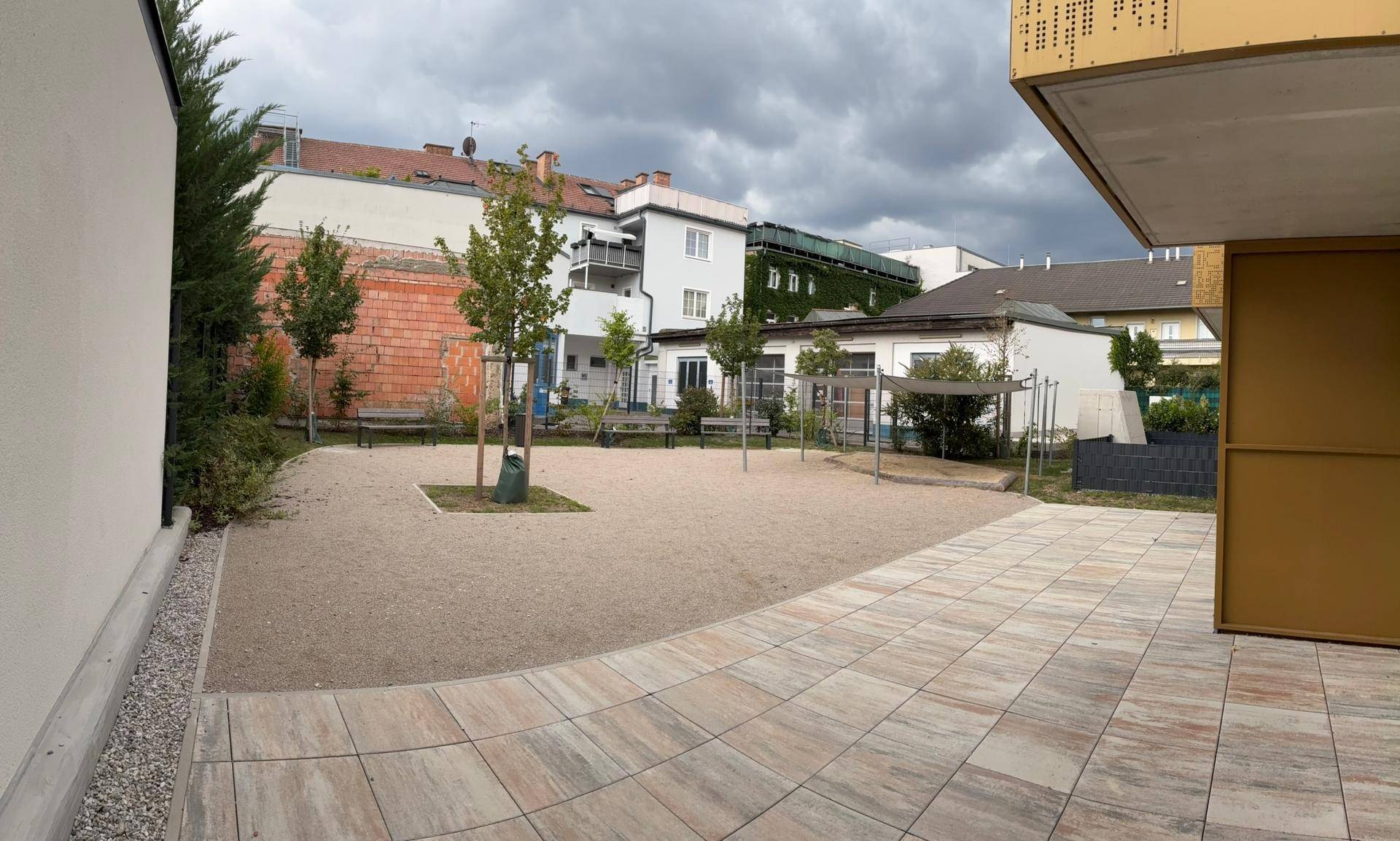 Außenbereich mit Sandspielplatz und Bäumen, umgeben von Wohngebäuden.