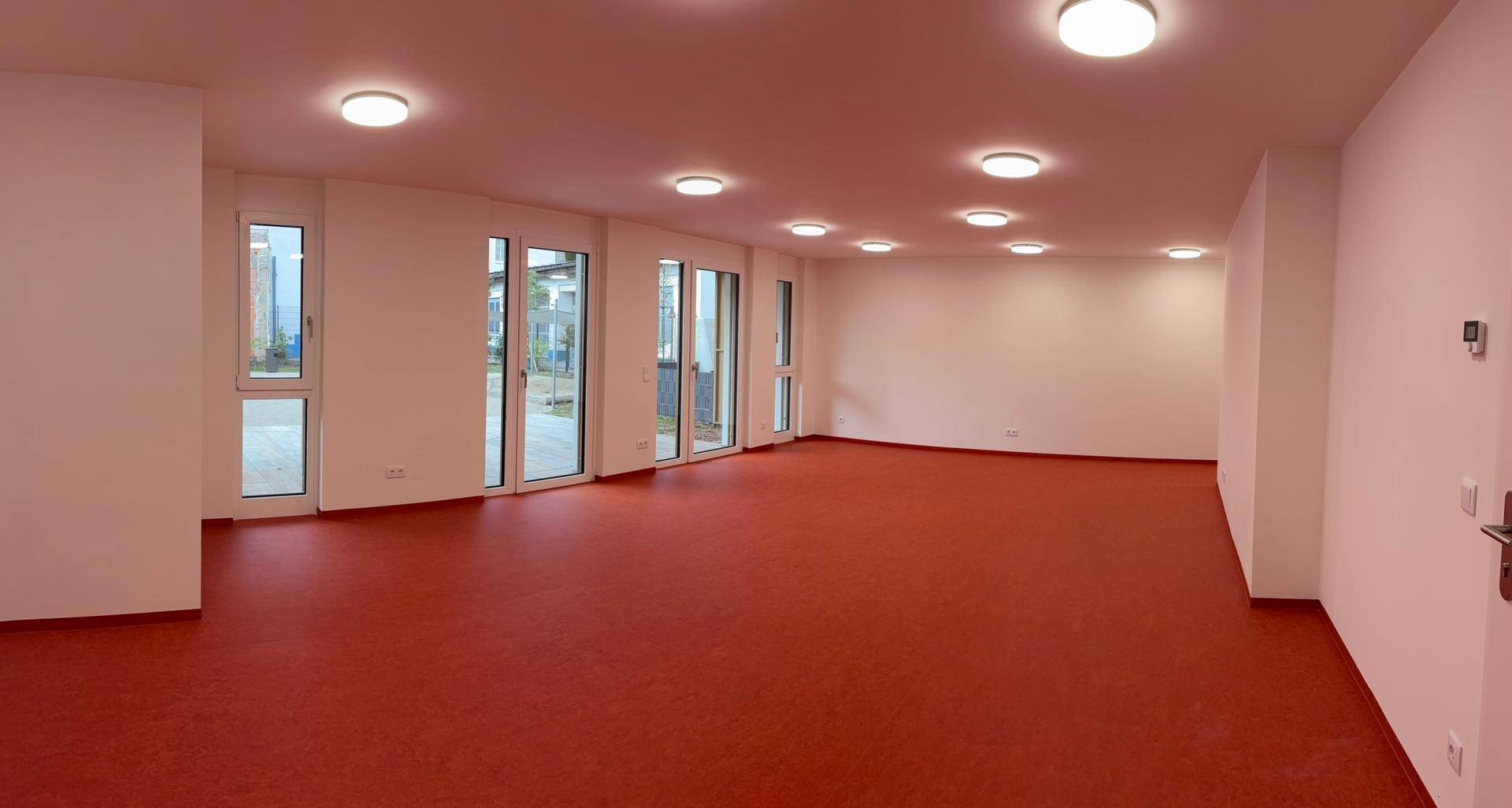Großer leerer Raum mit rotem Teppichboden und vielen Fenstern, die nach draußen führen.