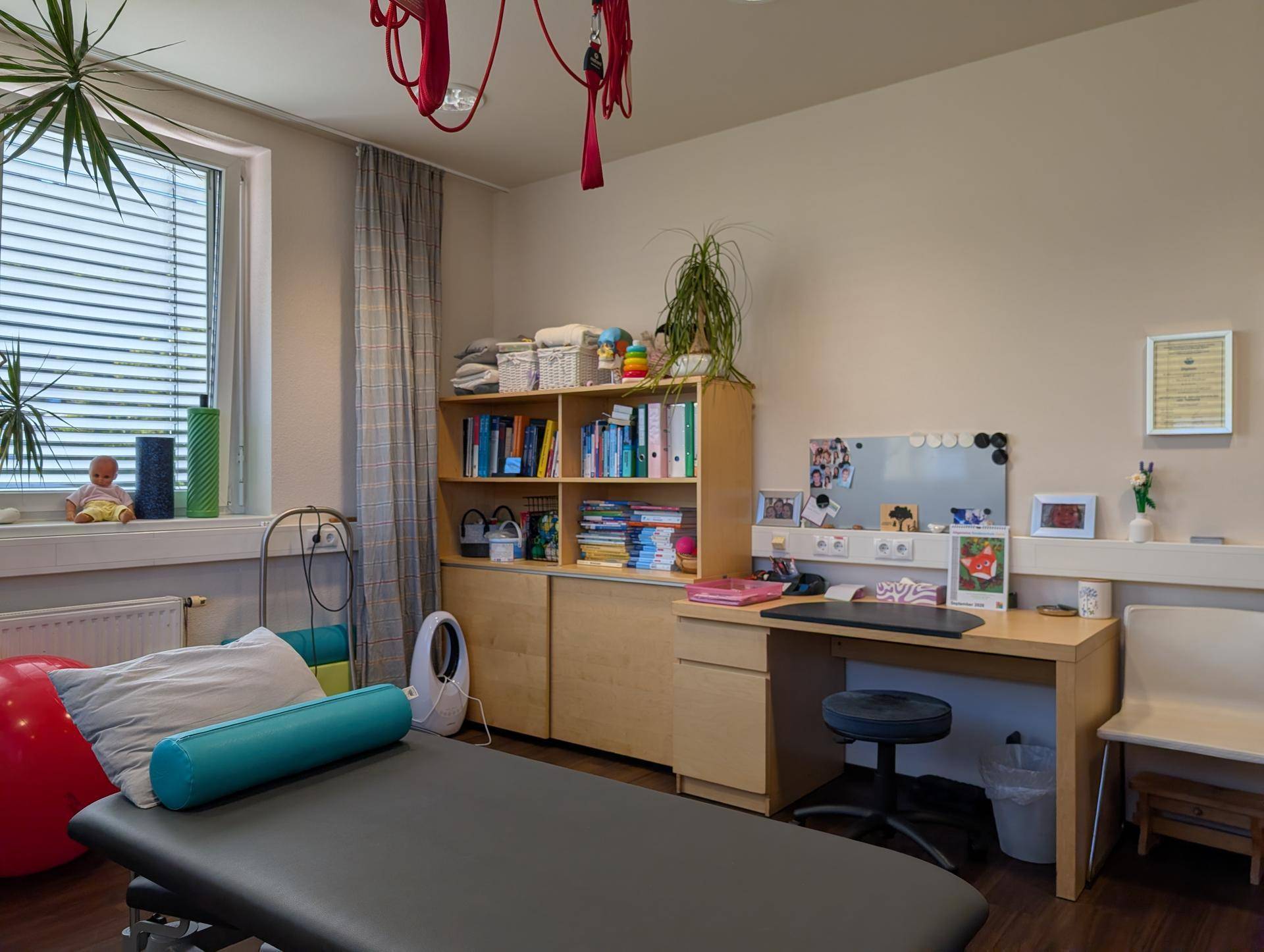 Therapieraum mit Schreibtisch, Bücherregal und Behandlungsliege, gut beleuchtet durch ein großes Fenster.