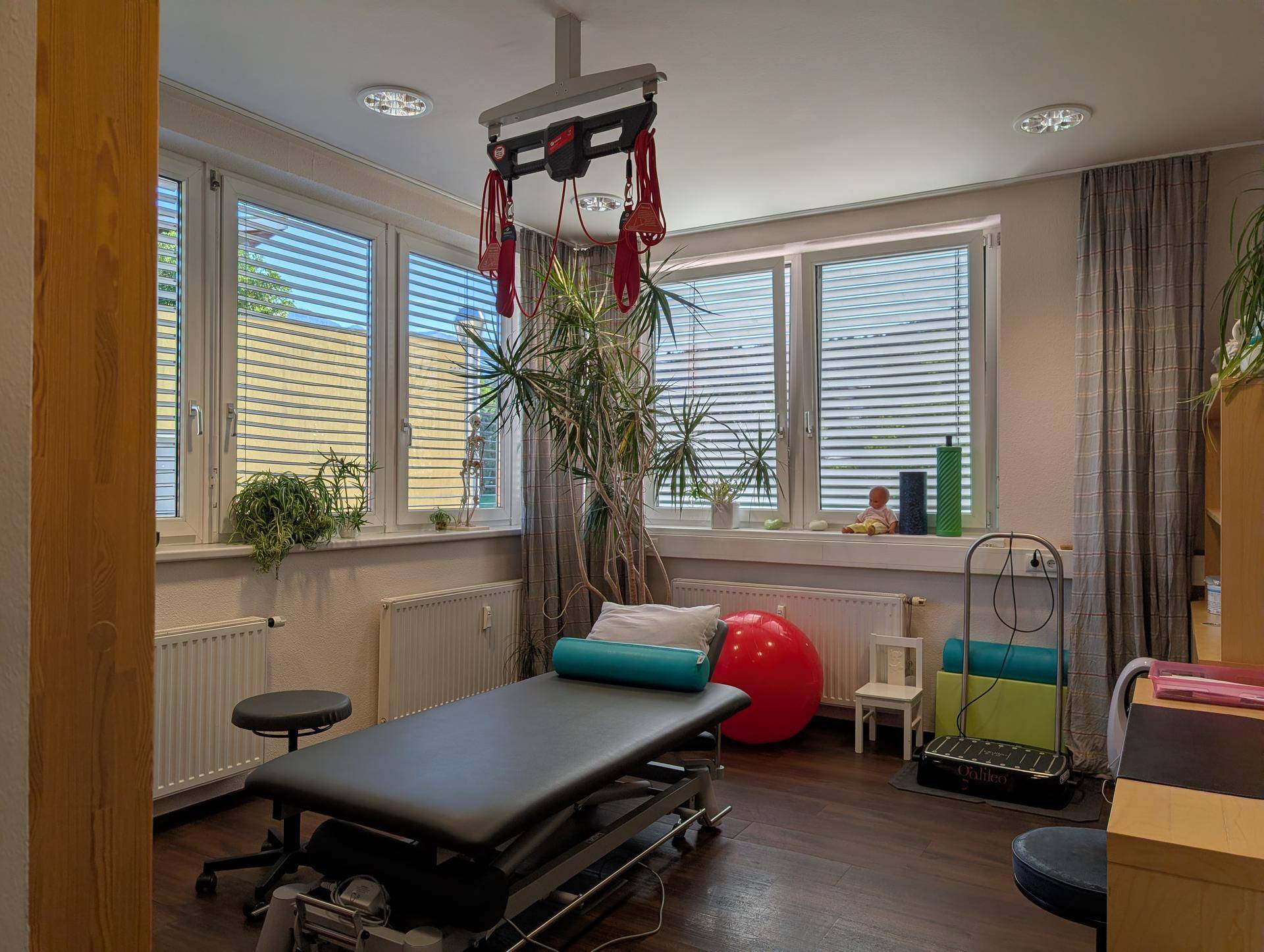 Heller Therapieraum mit Behandlungsliege, vielen Fenstern und einem Deckenlift für therapeutische Anwendungen.