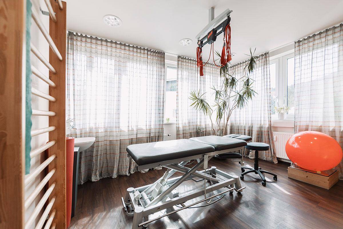 Therapieraum mit Behandlungsliege, Sprossenwand und großen Fenstern, die für viel Helligkeit sorgen.