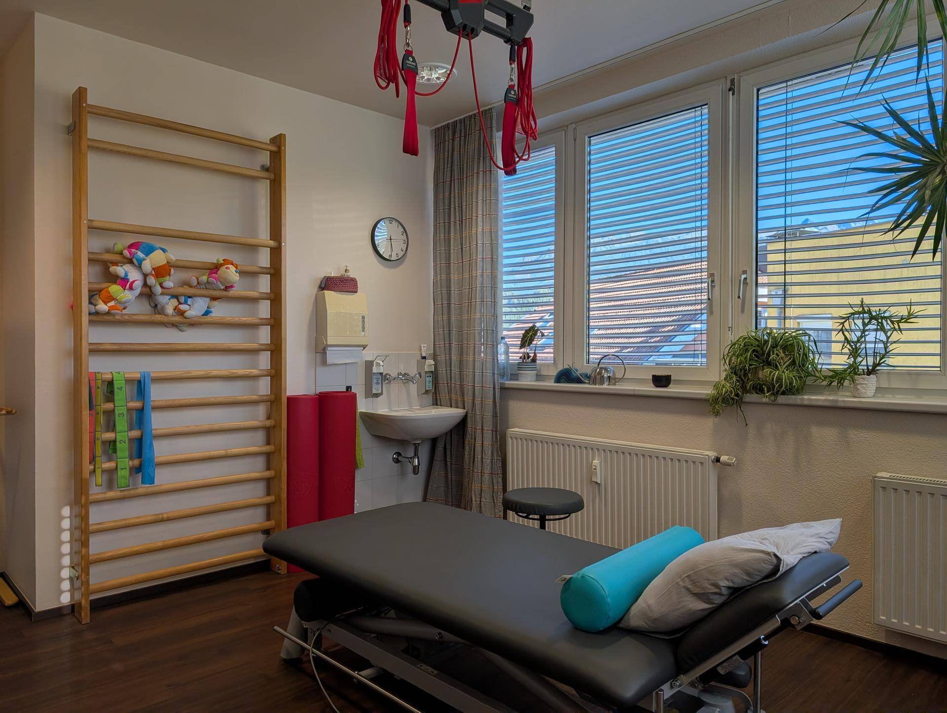 Therapieraum mit Behandlungsliege, Sprossenwand und Waschbecken, beleuchtet durch große Fenster mit Jalousien.