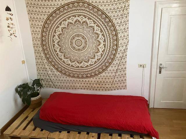 Schlafzimmer mit Palettenbett, roter Bettdecke und dekorativem Wandteppich.