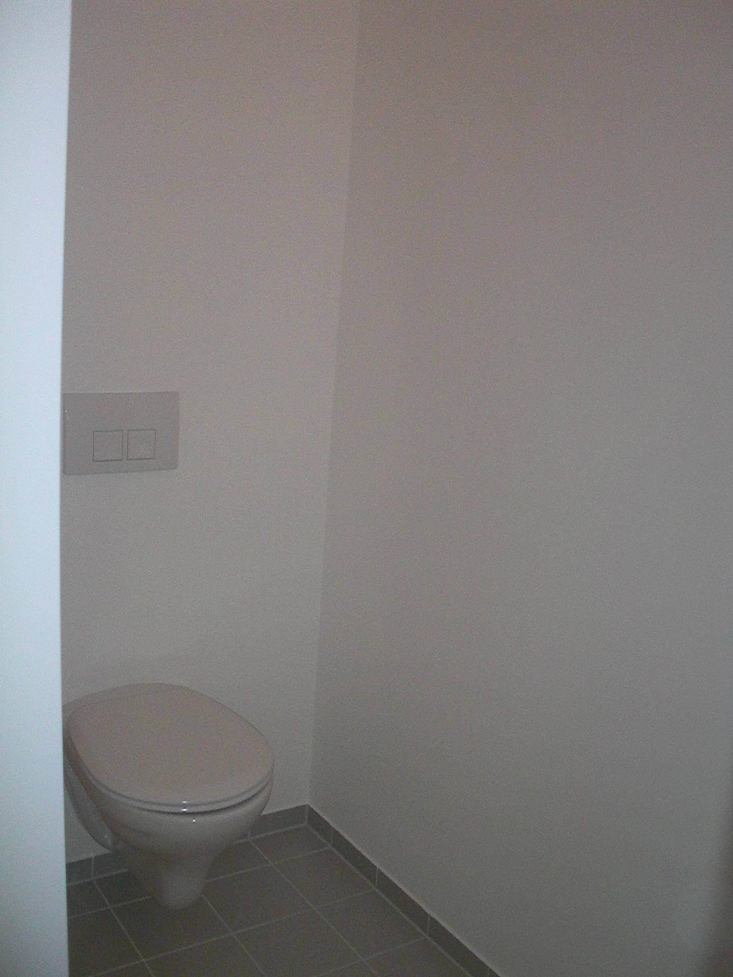 Separates WC mit wandhängender Toilette und grauen Bodenfliesen.